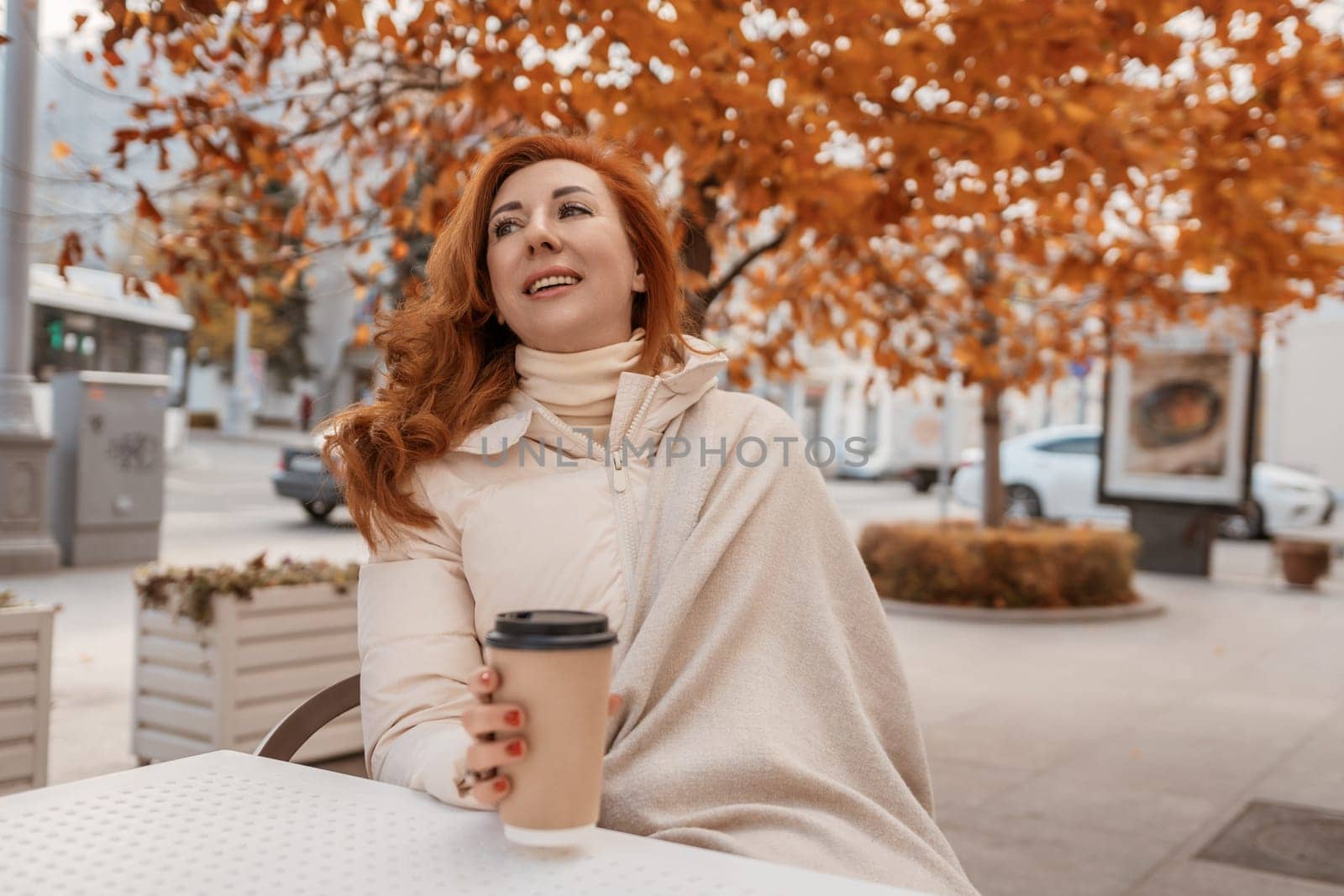 Redhead woman in beige coat sits outdoors at a cafe table, holding a disposable coffee cup. Autumn leaves frame the scene.