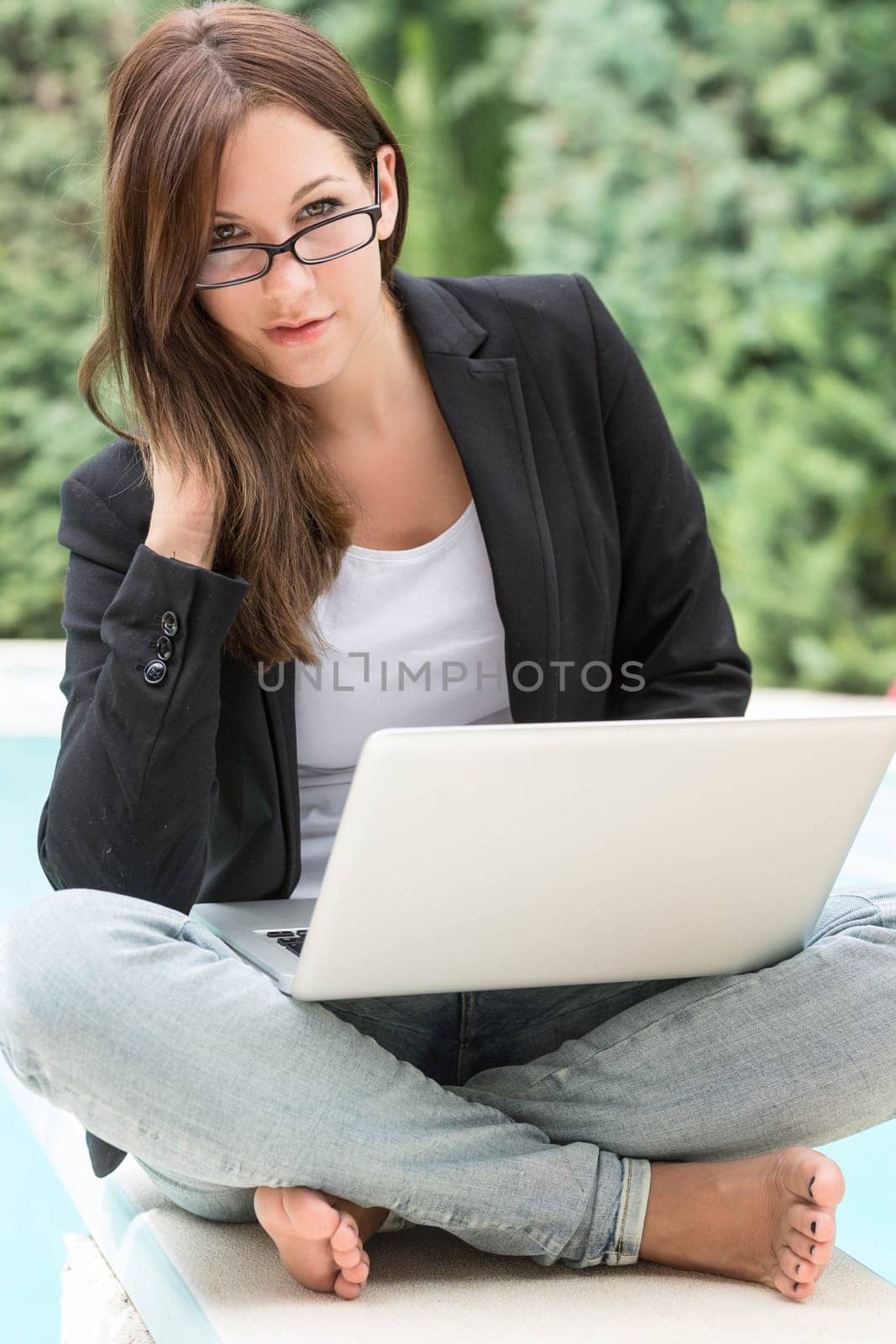 A young woman in blazer and glasses sits outside on the laptop