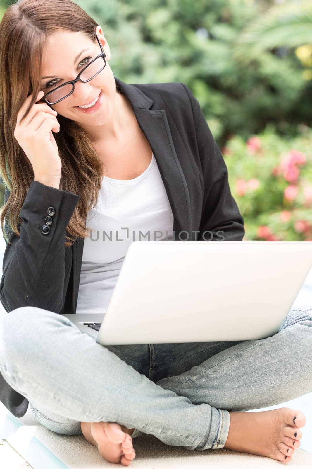 A young woman in blazer and glasses sits outside on the laptop