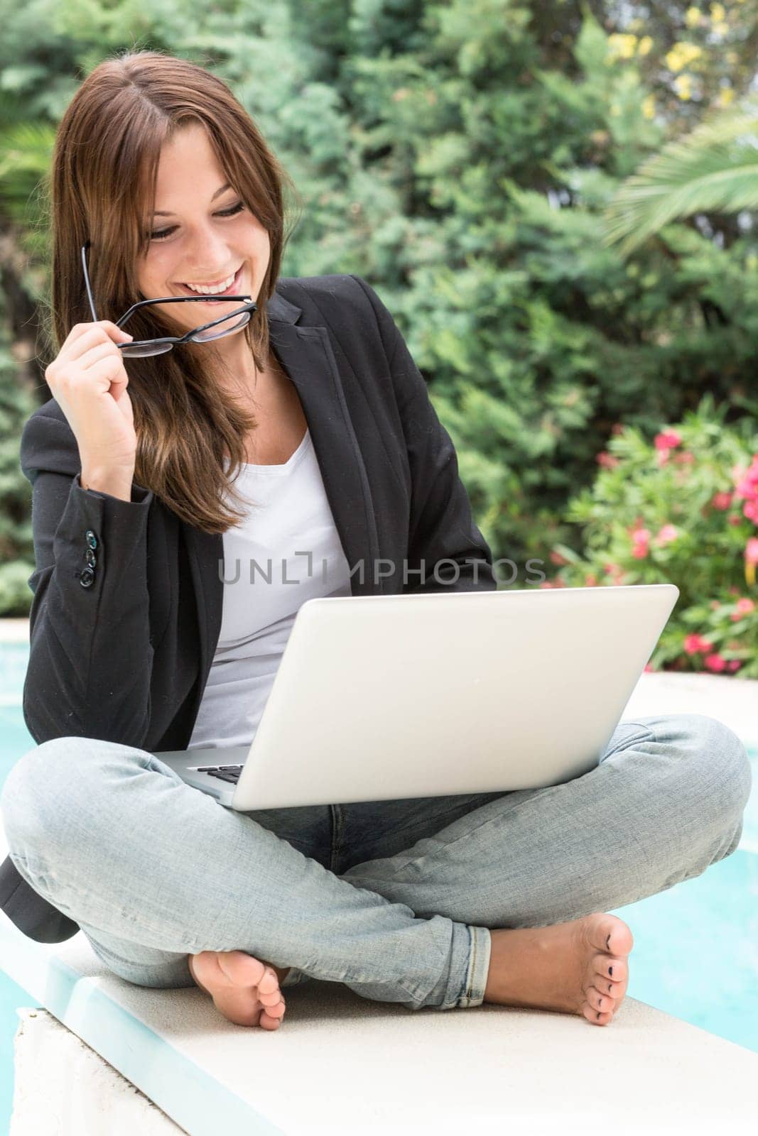 A young woman in blazer and glasses sits outside on the laptop