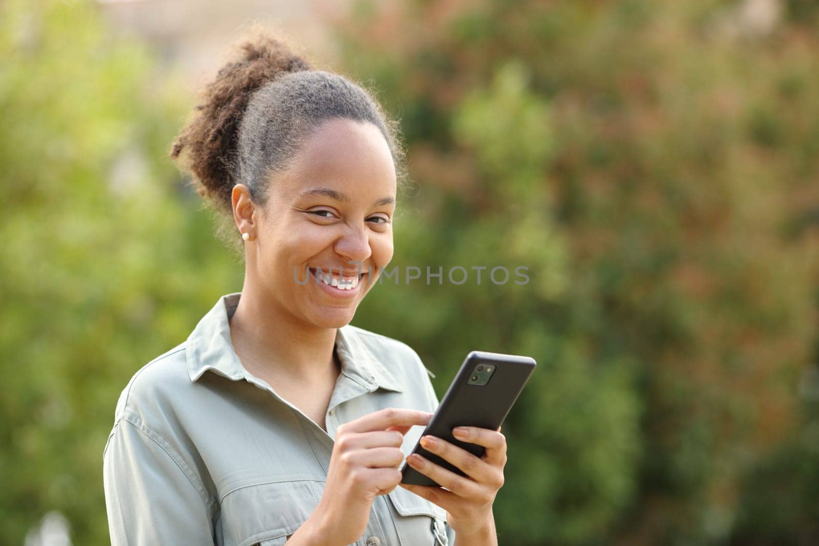 Happy black woman laughing holding phone looking at camera in a park