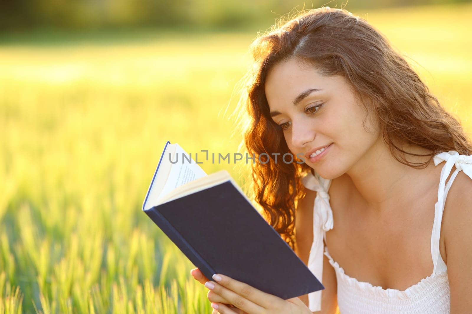 Happy woman sitting in a field reading a paper book at sunset