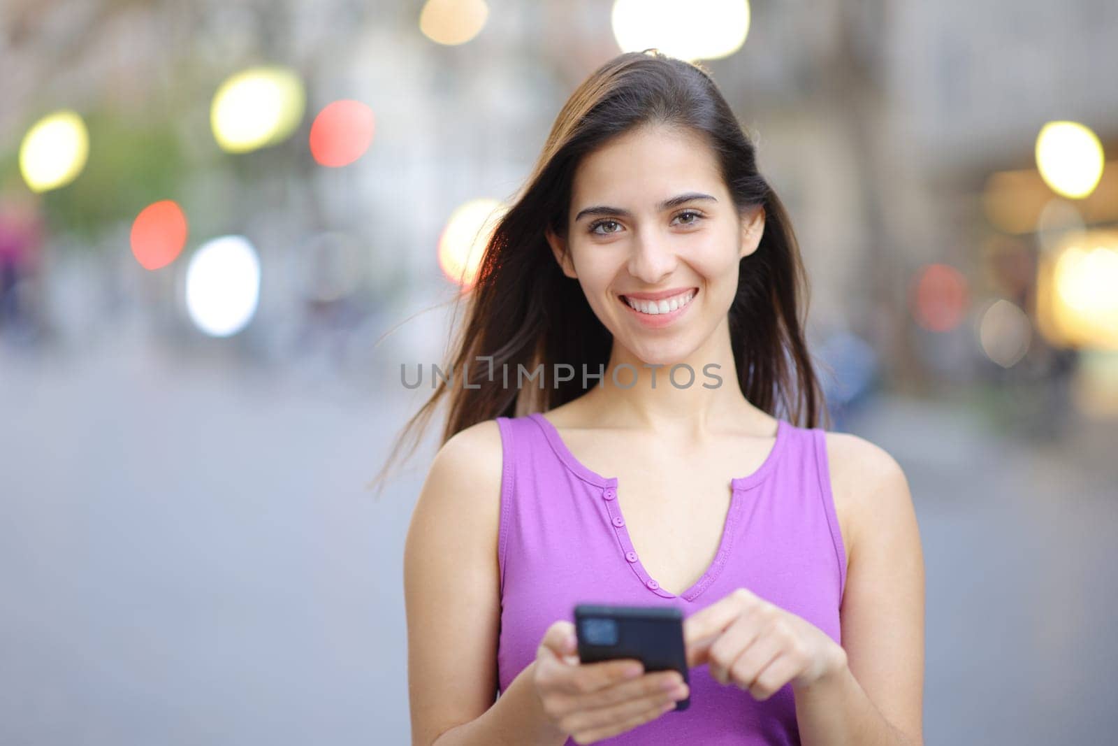 Front view portrait of a happy woman holding smart phone in the street looking at you