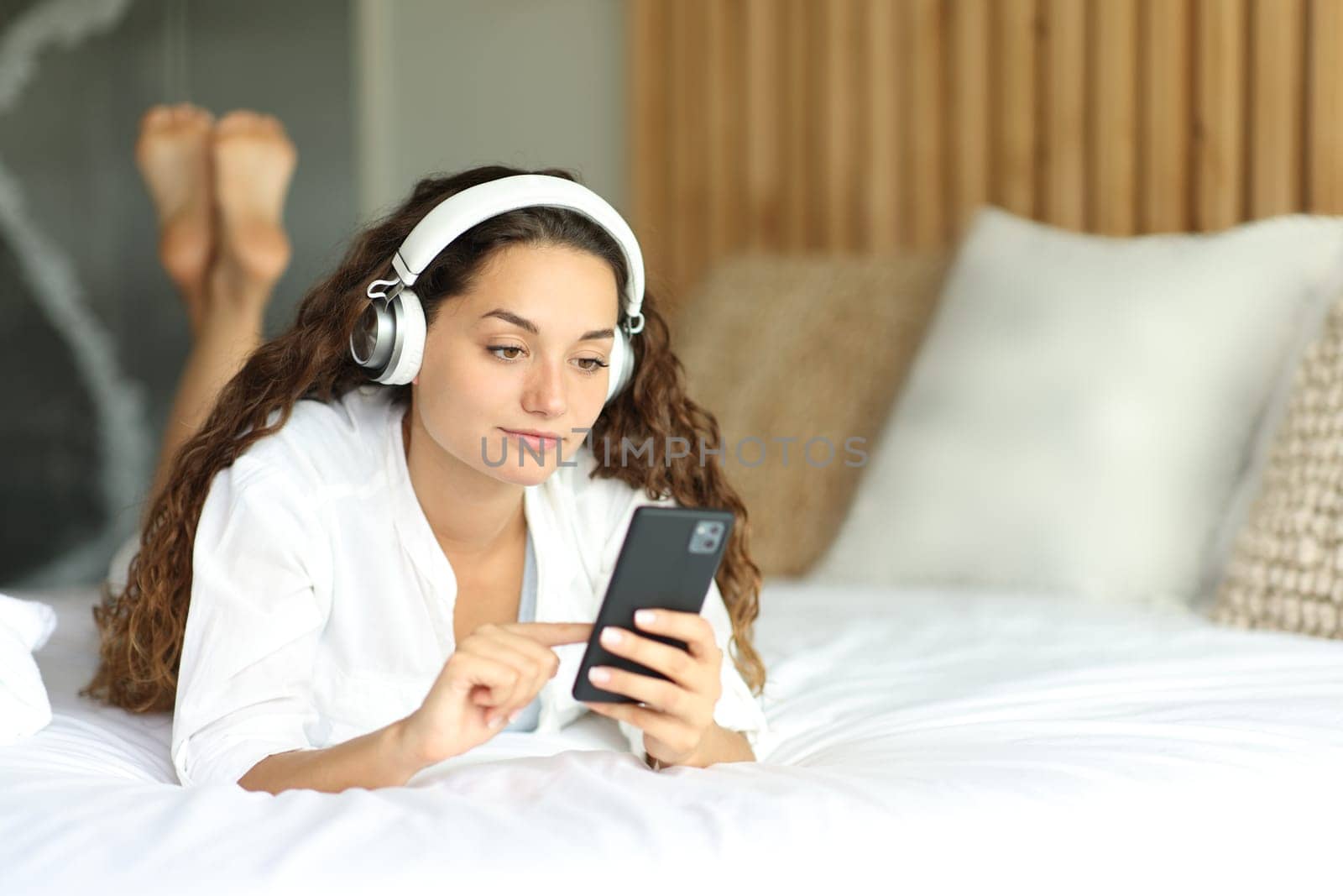 Woman with headphone on the bed watching media on phone lying on the bed in a room