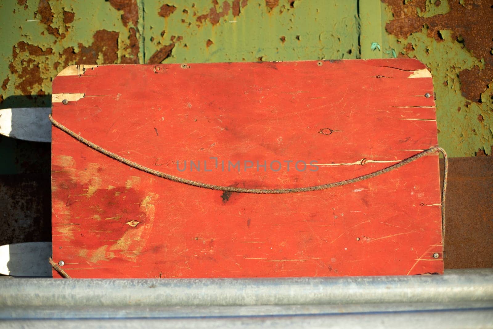 Old board is red against rusty wall. Composition of green and red color. Old thing in sunlight.