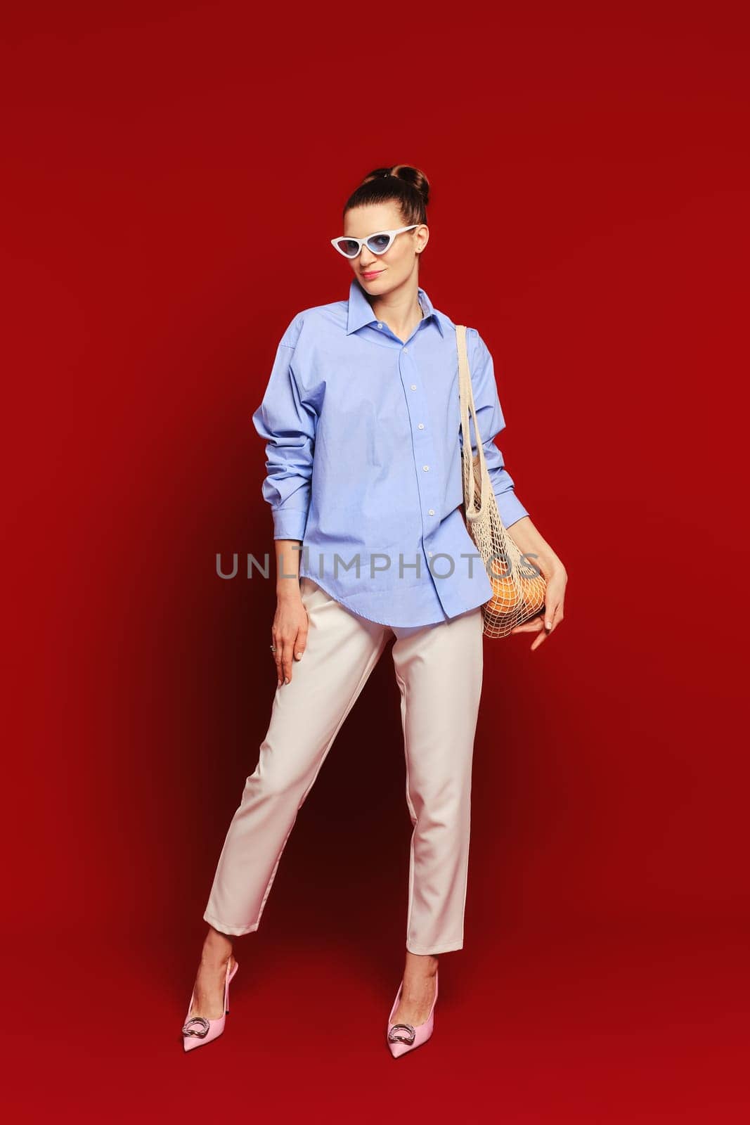 Cute woman in sunglasses with oranges in reusable string bag over her shoulder on ruby red background. Concept of sustainable living and care of the environment.