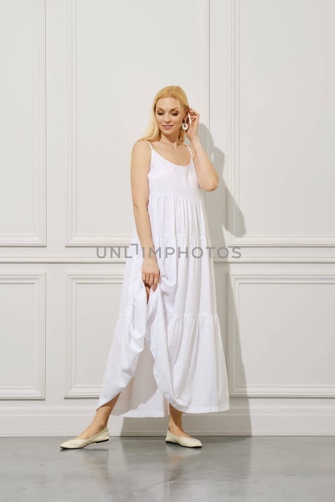 Cute and tender adult woman lower her eyes as like she is shy, she touching her long blonde hair and smile
