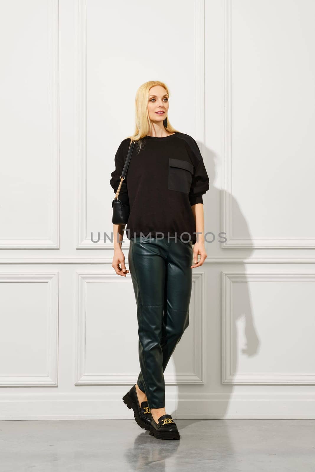 Cheerful woman stands in leather trousers and pullover in bright room. From head to toe she is dressed casually but fashionably with their footwear completing the look.