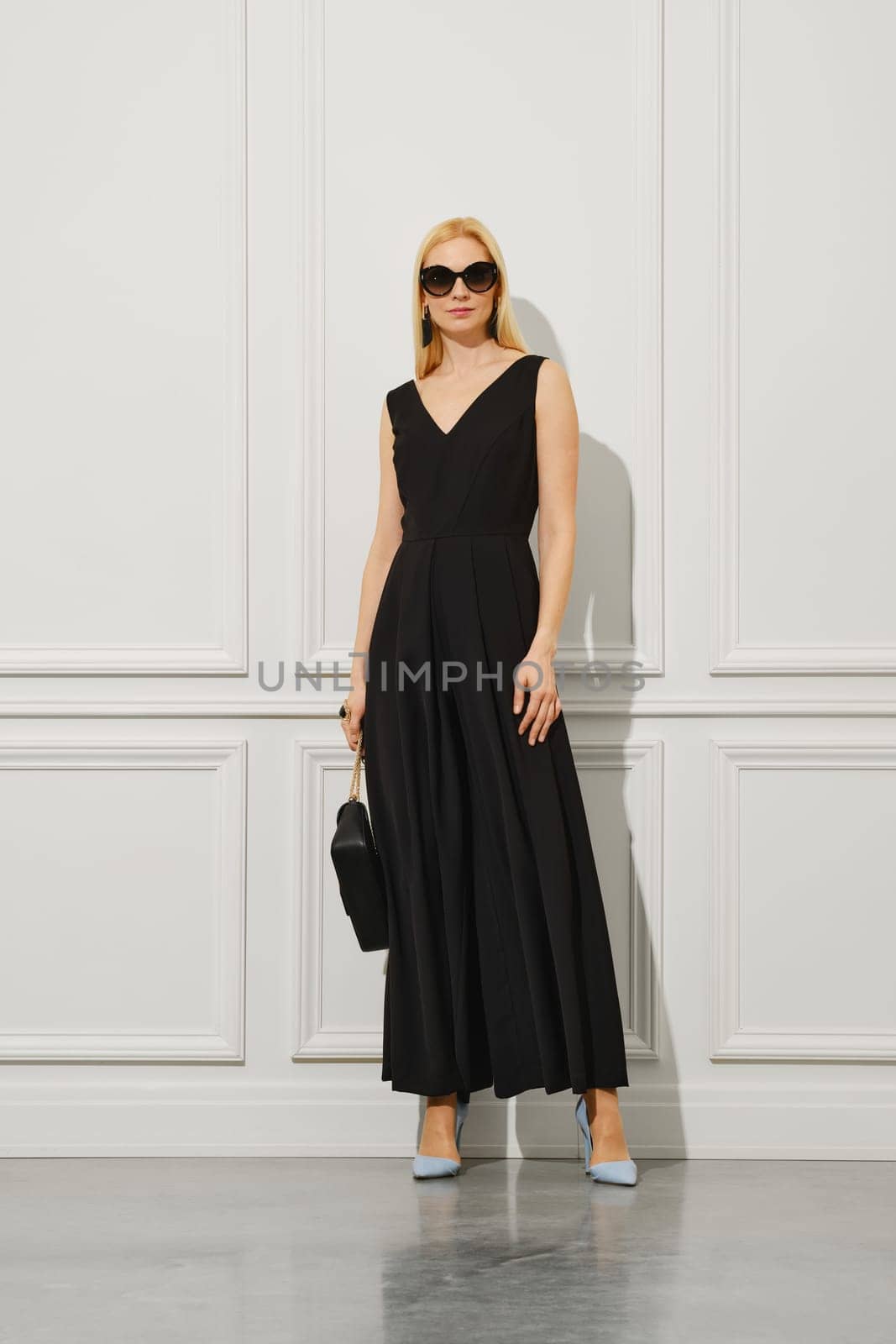 Confident businesswoman in long black dress and sunglasses stands in a bright room holding little handbag