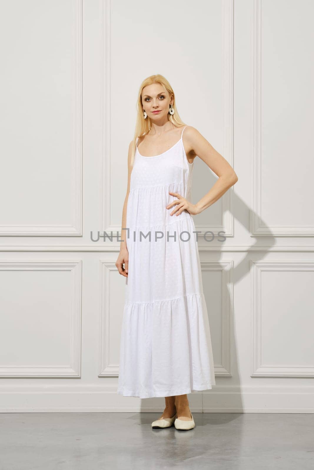 A beautiful woman with long, blond hair in a white sundress stands indoors and looks into the camera