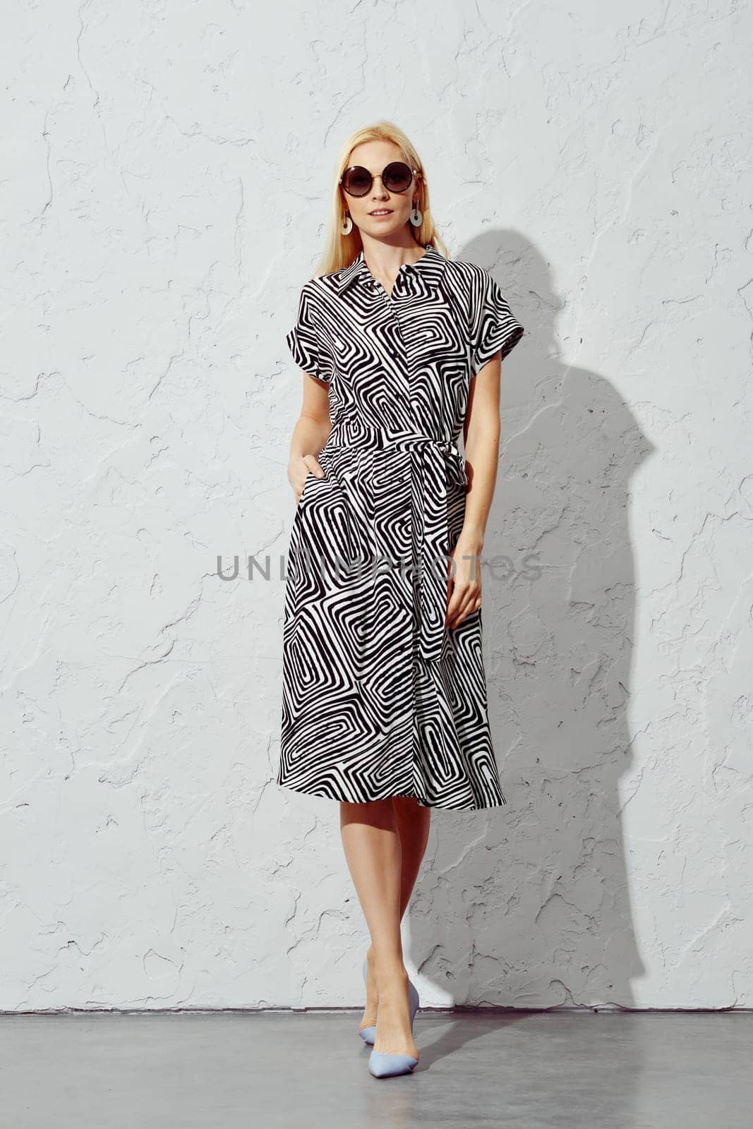 A stylish woman walking forward in a patterned dress and sunglasses under harsh sunlight, looking directly at the camera