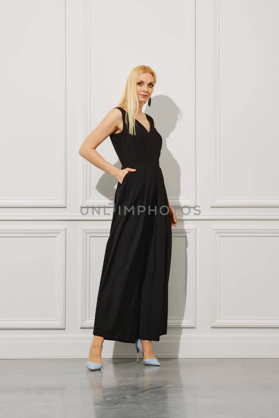 A blond businesswoman stands in a bright room, dressed fashionably in long black dress and wearing stilettos. She holds one hand in a pocket