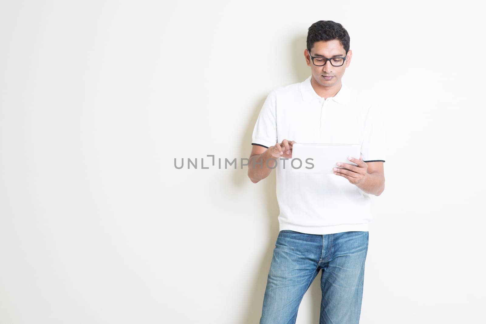 Portrait of handsome Indian guy using tablet pc, standing on plain background with shadow, copy space at side.