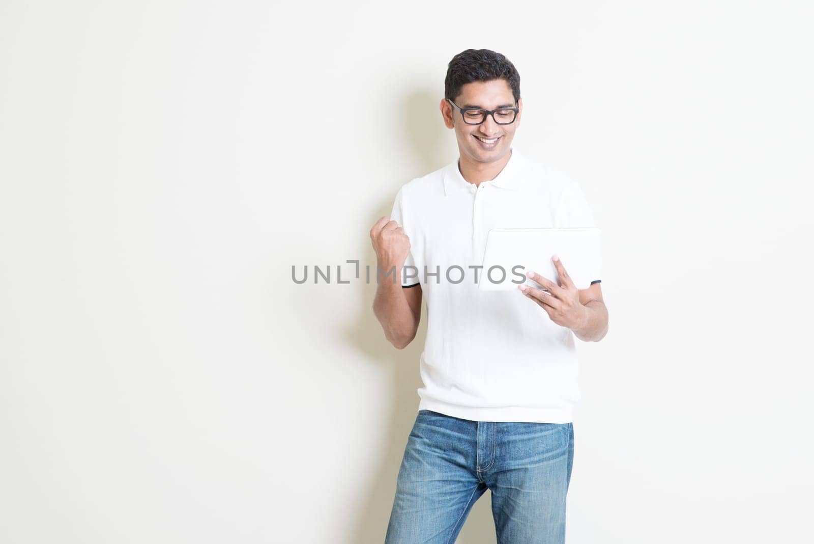 Portrait of excited Indian guy using tablet pc and celebrate his success . Asian man standing on plain background with shadow and copy space. Handsome male model.