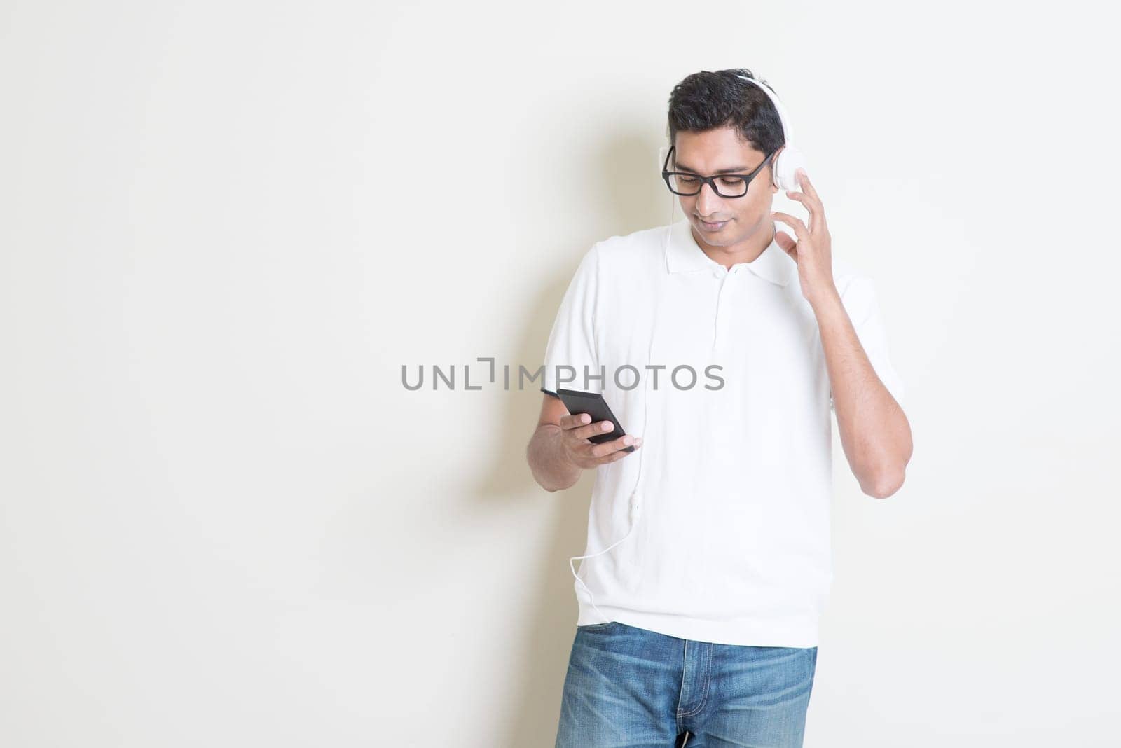 Smartphone social media concept. Indian guy using mobile phone. Asian man relaxed and listening music with headset, standing on plain background. Handsome male model.
