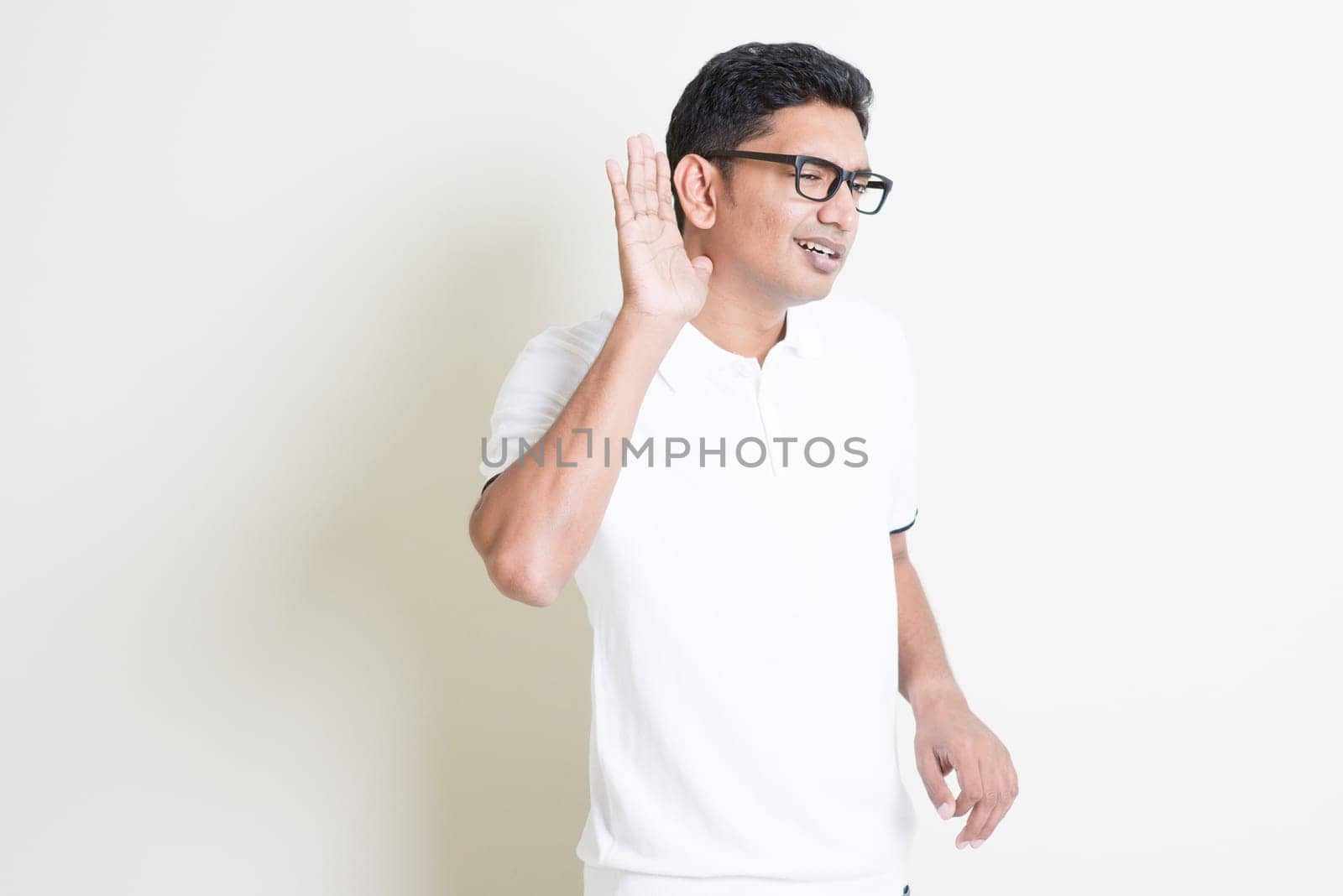 Portrait of Indian guy listening, hand cupped beside ears. Asian man standing on plain background with shadow and copy space. Handsome male model.