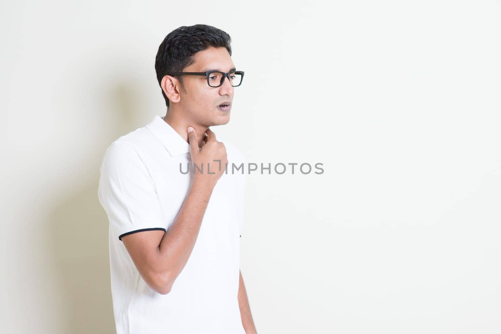 Portrait of Indian guy sore throat, hand on neck clearing throat. Asian man standing on plain background with shadow and copy space. Handsome male model.