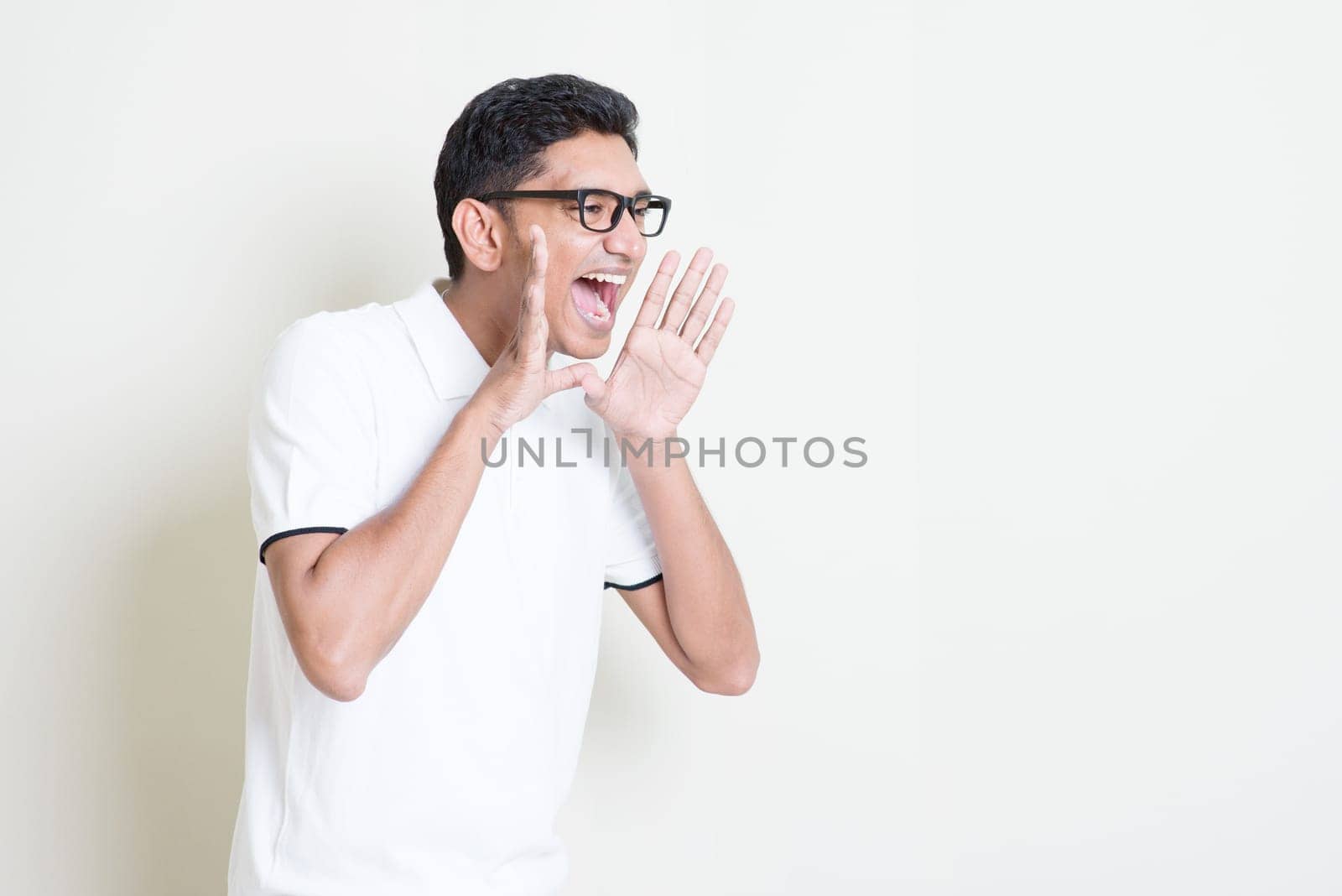 Portrait of Indian guy shouting loud, mouth open and hand cupped beside. Asian man standing on plain background with shadow and copy space. Handsome male model.
