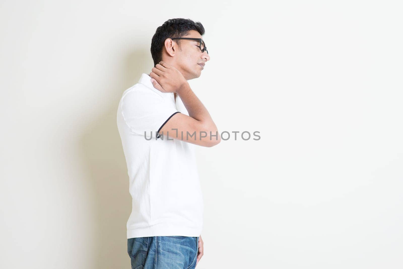 Portrait of Indian guy neck pain, massaging with hand. Asian man standing on plain background with shadow and copy space. Handsome male model.