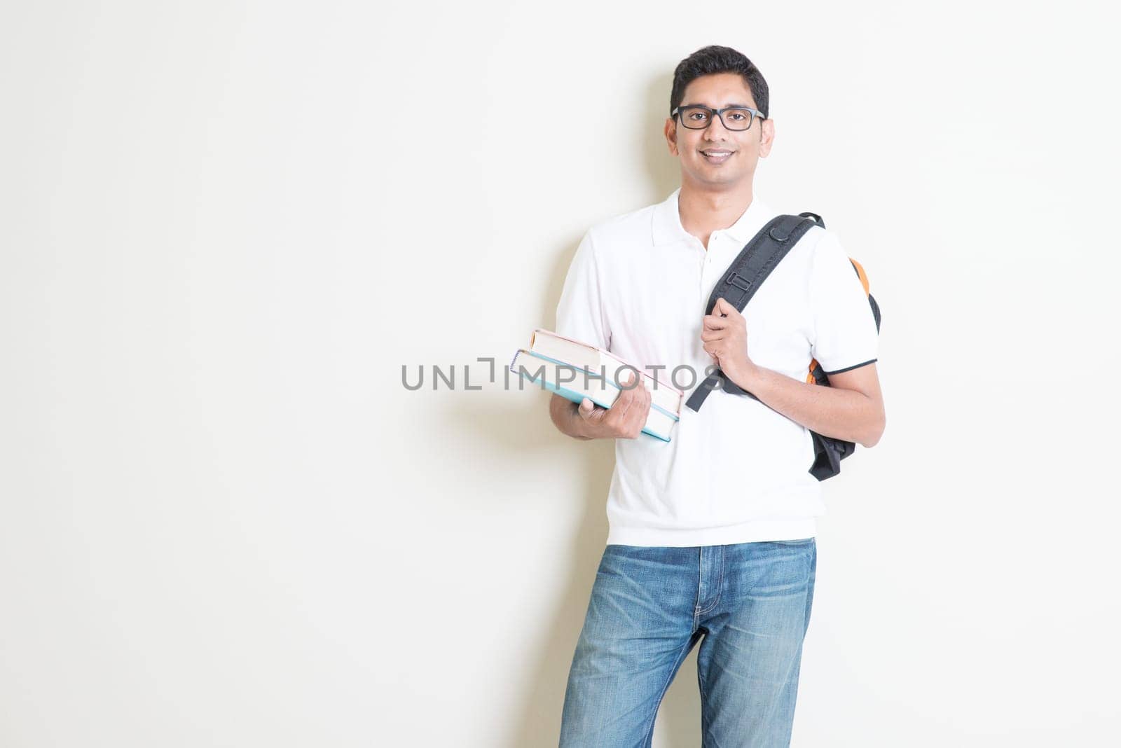 Portrait of adult Indian university student with books and bag. Asian man standing on plain background with shadow and copy space. Handsome mixed race male model.