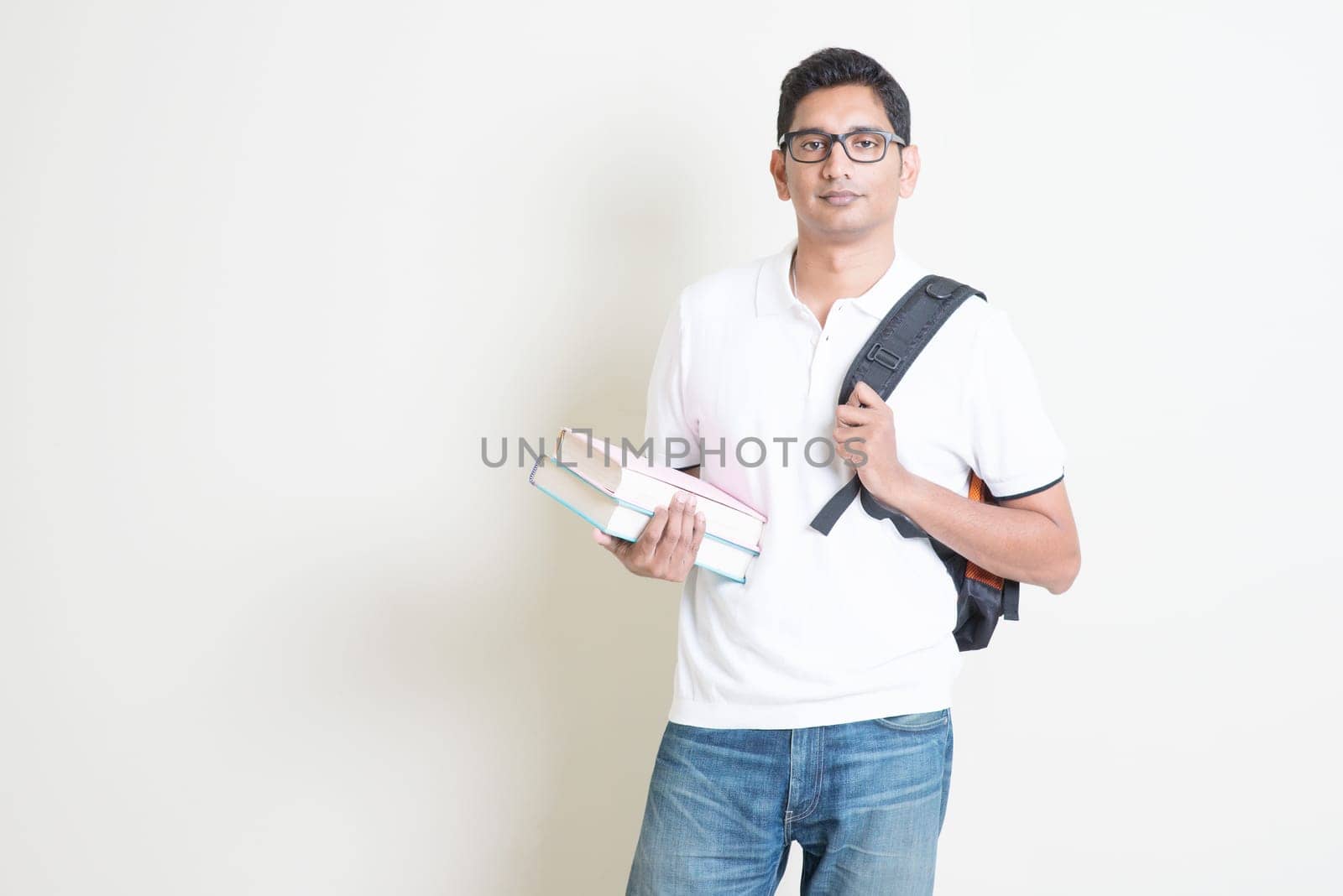 Portrait of Indian college student with books and backpack. Asian man standing on plain background with shadow and copy space. Handsome mixed race male model.