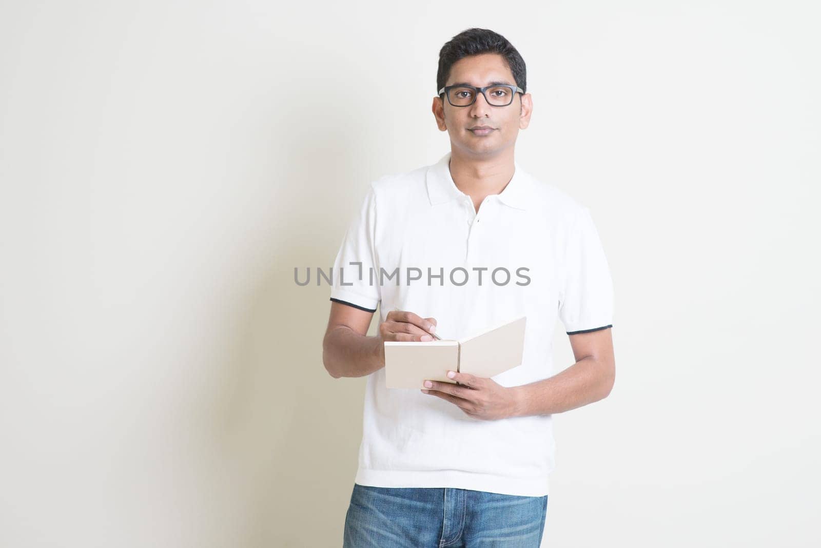 Portrait of Indian guy taking note on booklet. Asian man standing on plain background with shadow and copy space.