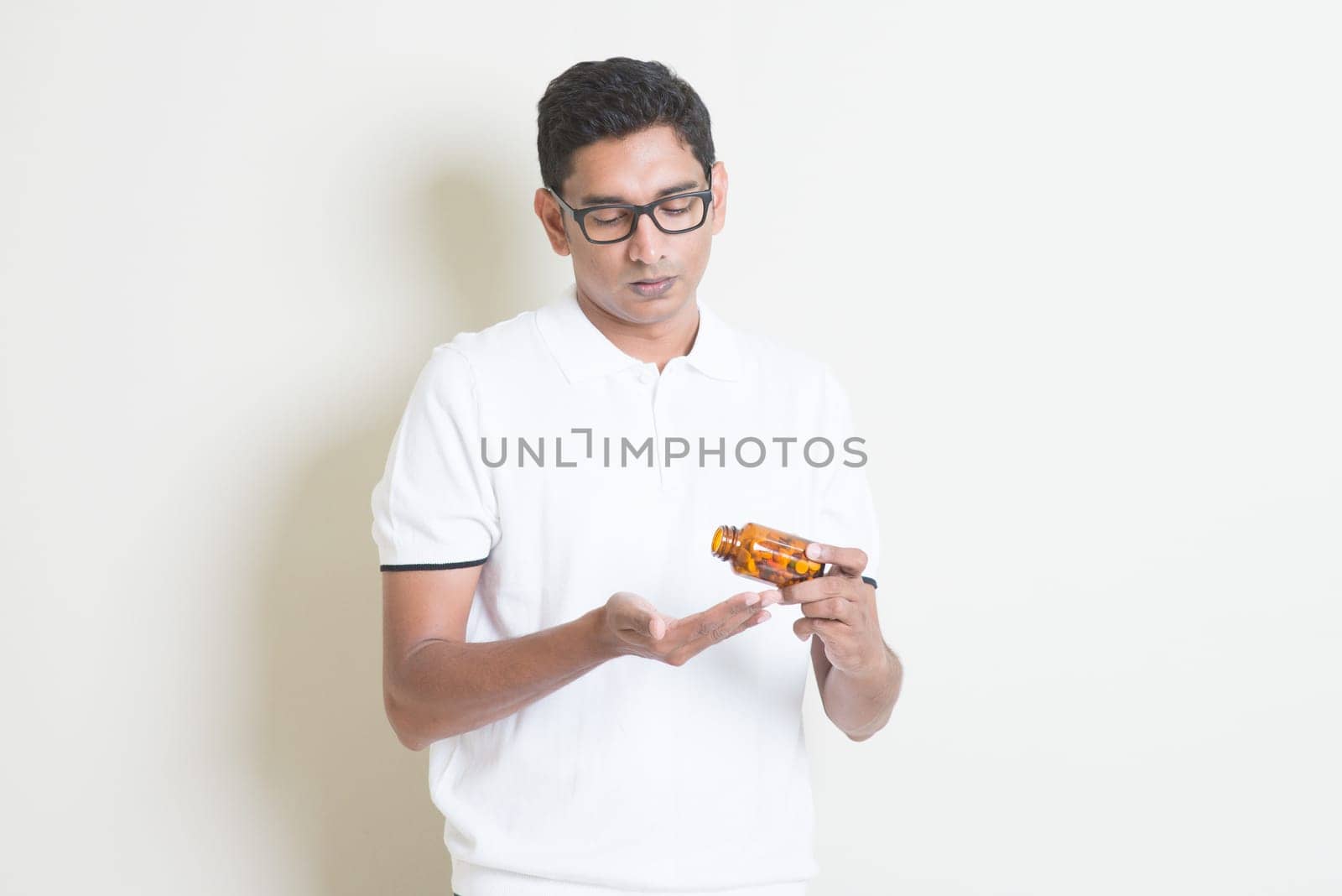 Sick Indian guy taking tablet medicine. Asian man standing on plain background with shadow and copy space.