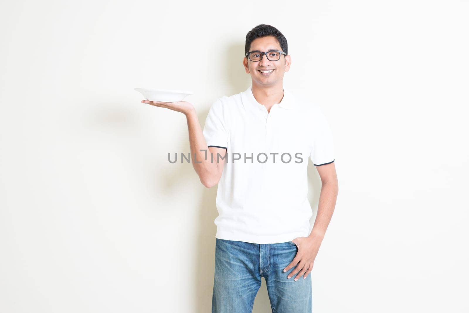 Dining concept. Indian waiter holding an empty plate on hand, ready for food. Asian man standing on plain background with shadow and copy space. Handsome male model.