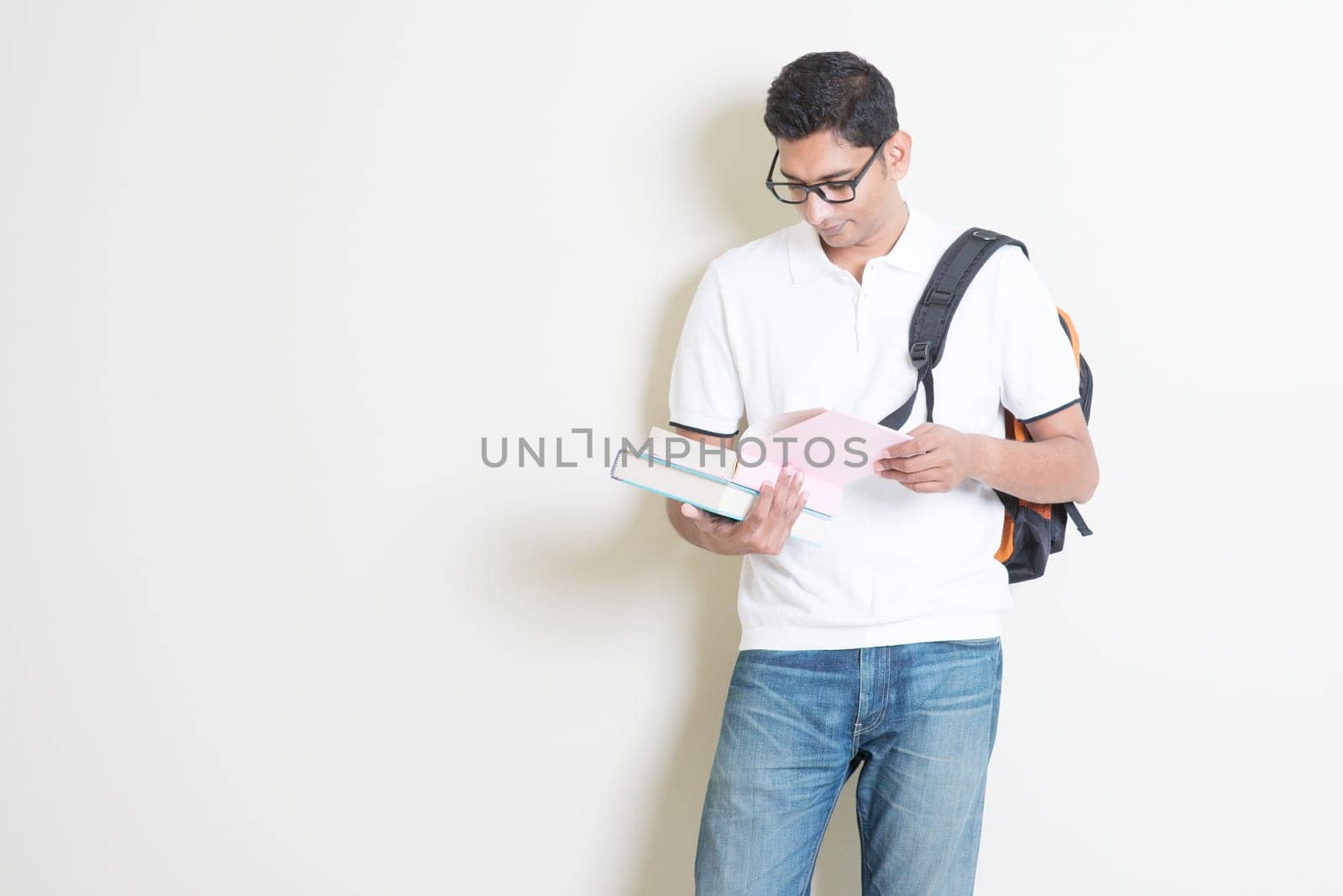 Portrait of adult Indian university student with bag reading book. Asian man standing on plain background with shadow and copy space. Handsome mixed race male model.