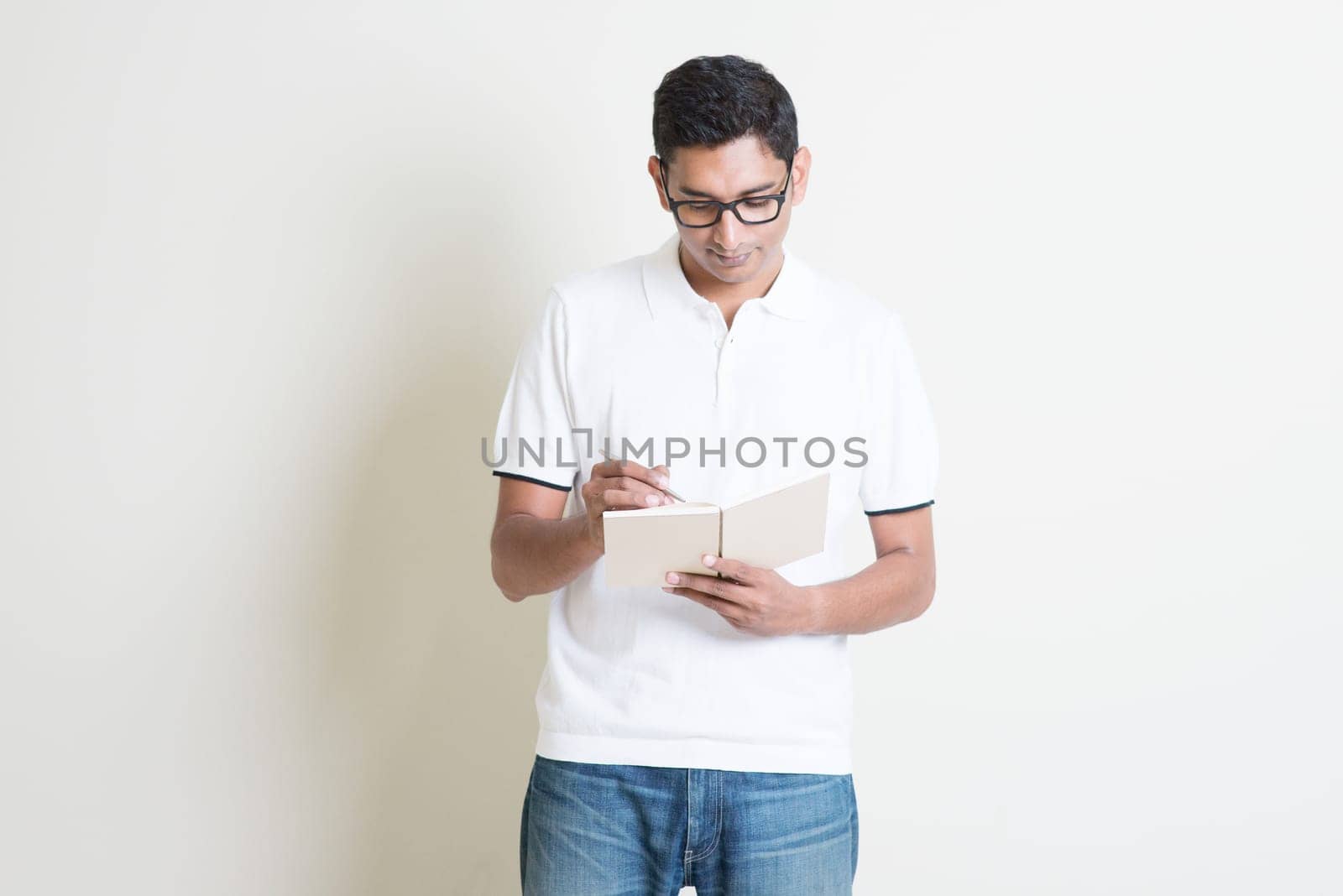 Portrait of Indian guy taking note on book. Asian man standing on plain background with shadow and copy space. Handsome male model.