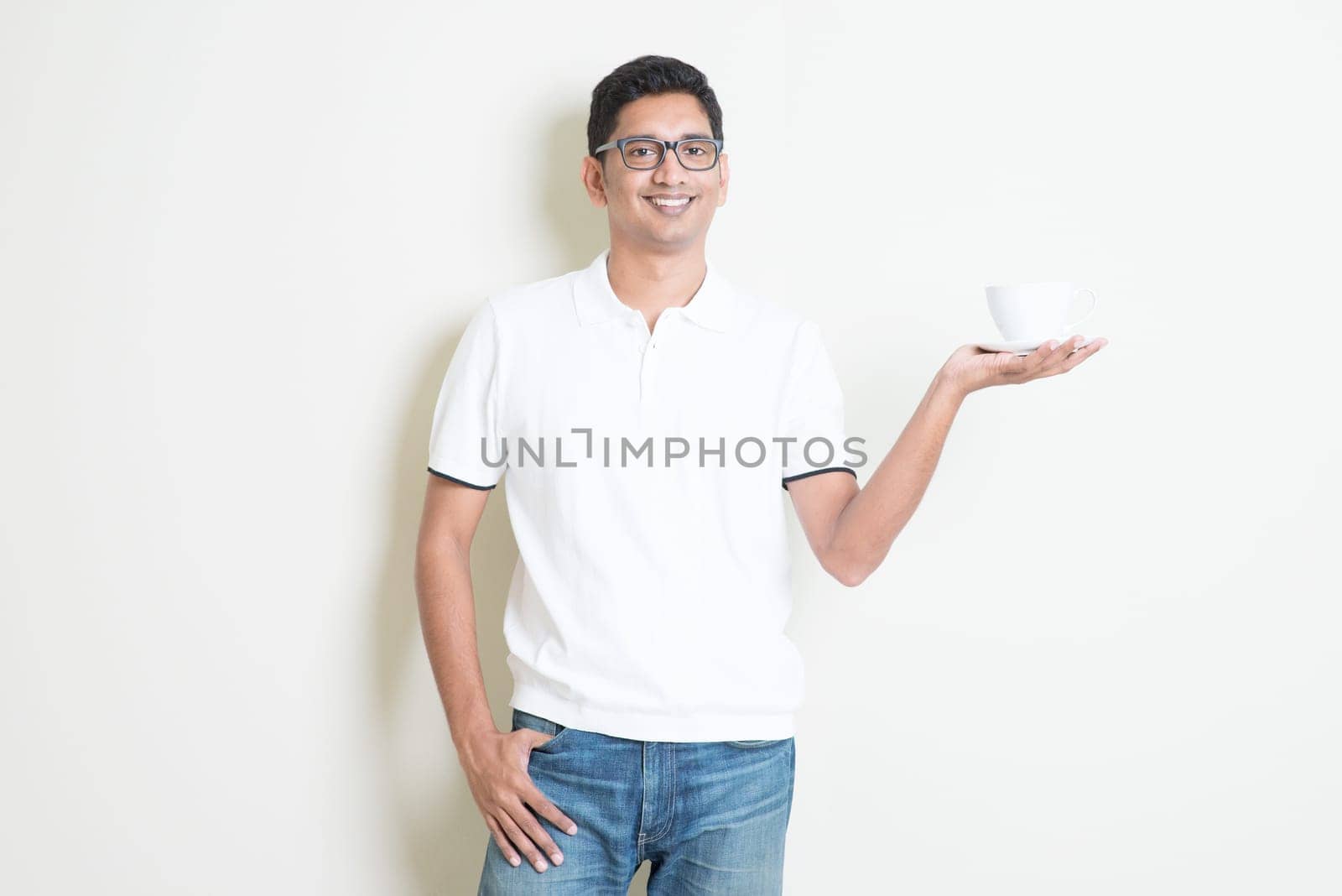 Indian guy holding a cup of coffee on hand. Asian man standing on plain background with shadow and copy space. Handsome male model.