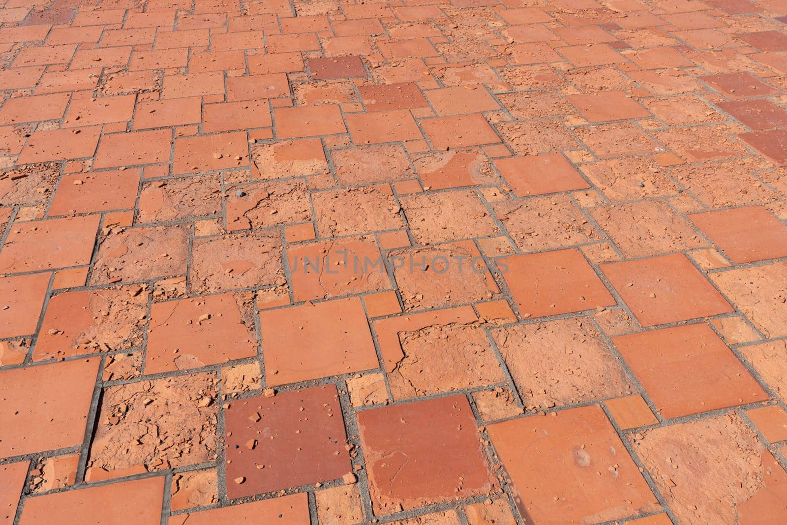 Red square floor tiles damaged by weathering or material defects. The surface is crumbling and the plates are disintegrating.