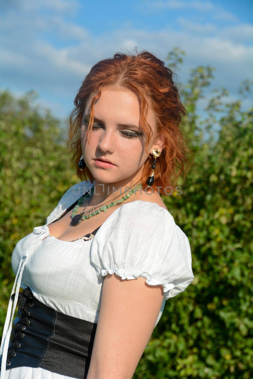 Portrait of a pretty young woman wearing white top, looking down, standing outdoors with green trees and blue sky