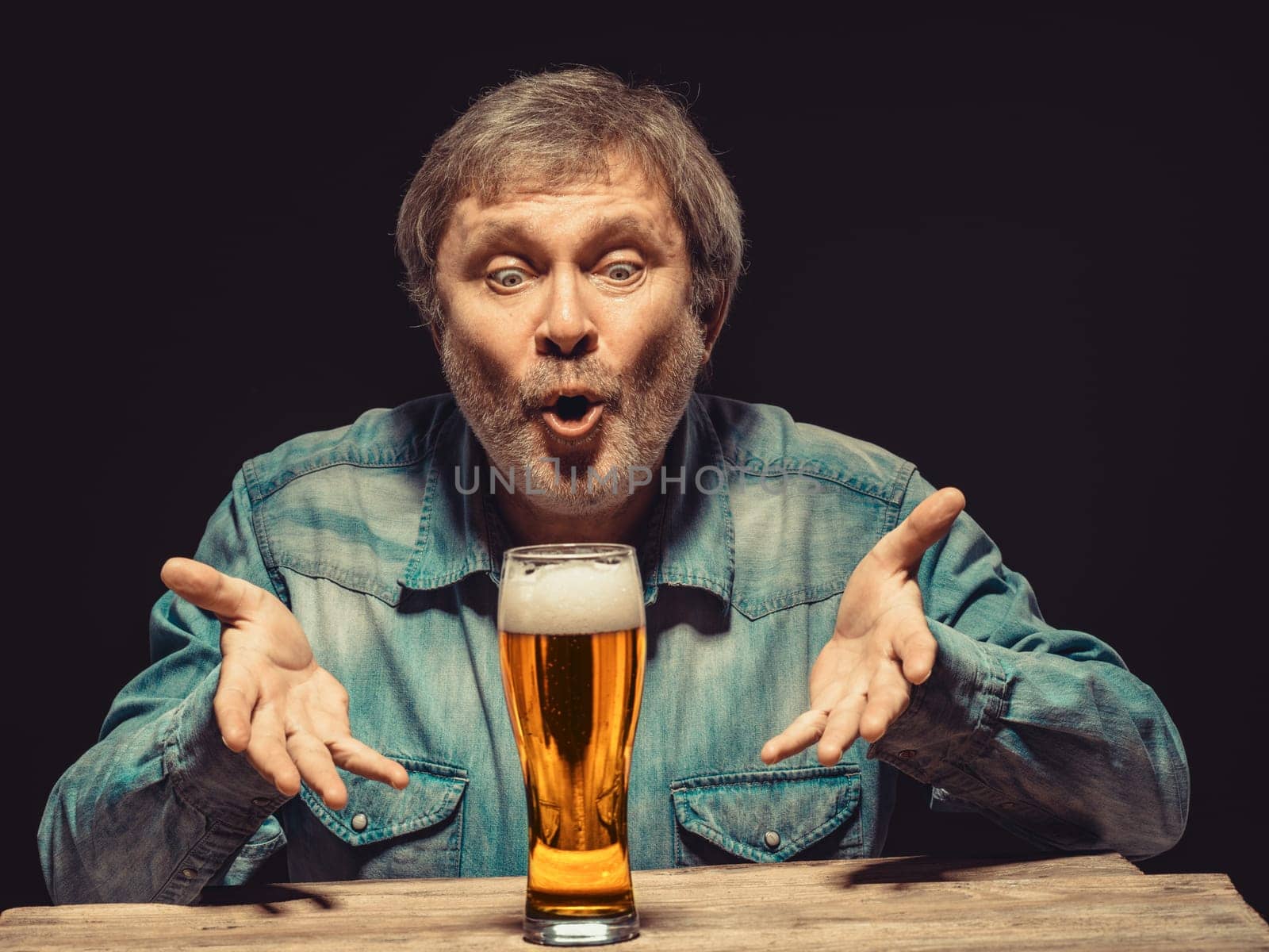 Enjoying his favorite beer.  The front view of handsome spellbound man in denim shirt with glass of beer, sitting at the wooden table. Concept of desire and love for beer