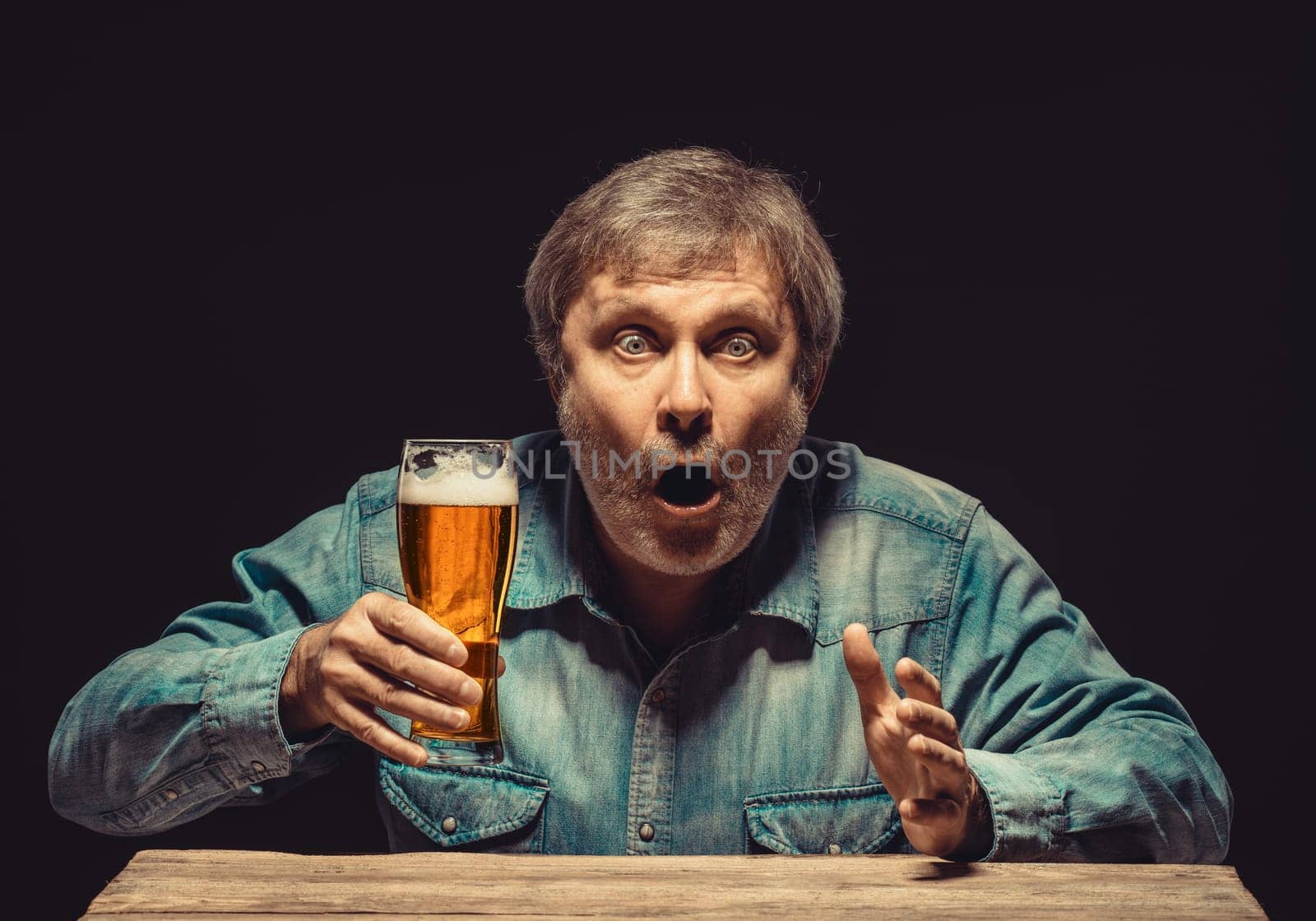 The front view of handsome man as enchanted and emotional fan in denim shirt with glass of beer, sitting at the wooden table. Concept of  surprise and exciting spectacle