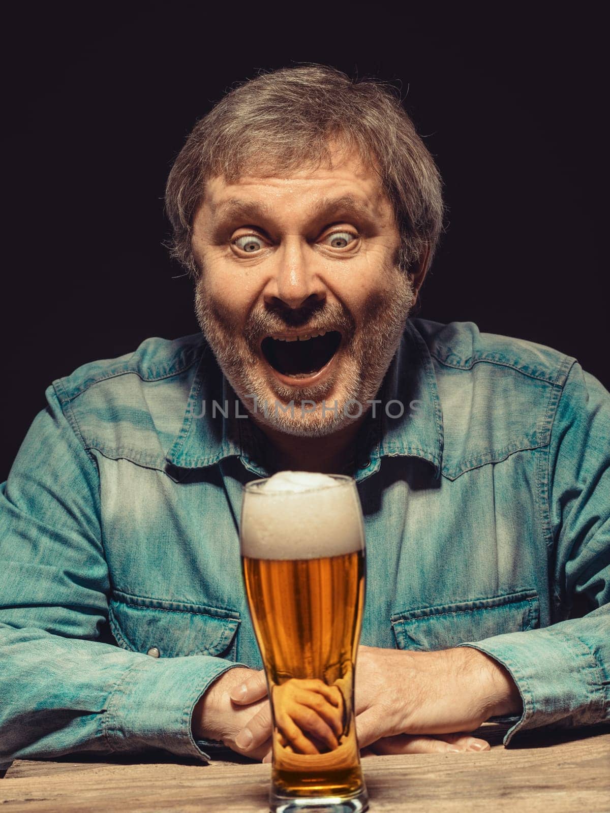 Enjoying his favorite beer.  The front view of handsome screaming man as fan in denim shirt with glass of beer, sitting at the wooden table. Concept of enthusiasm and ecstasy