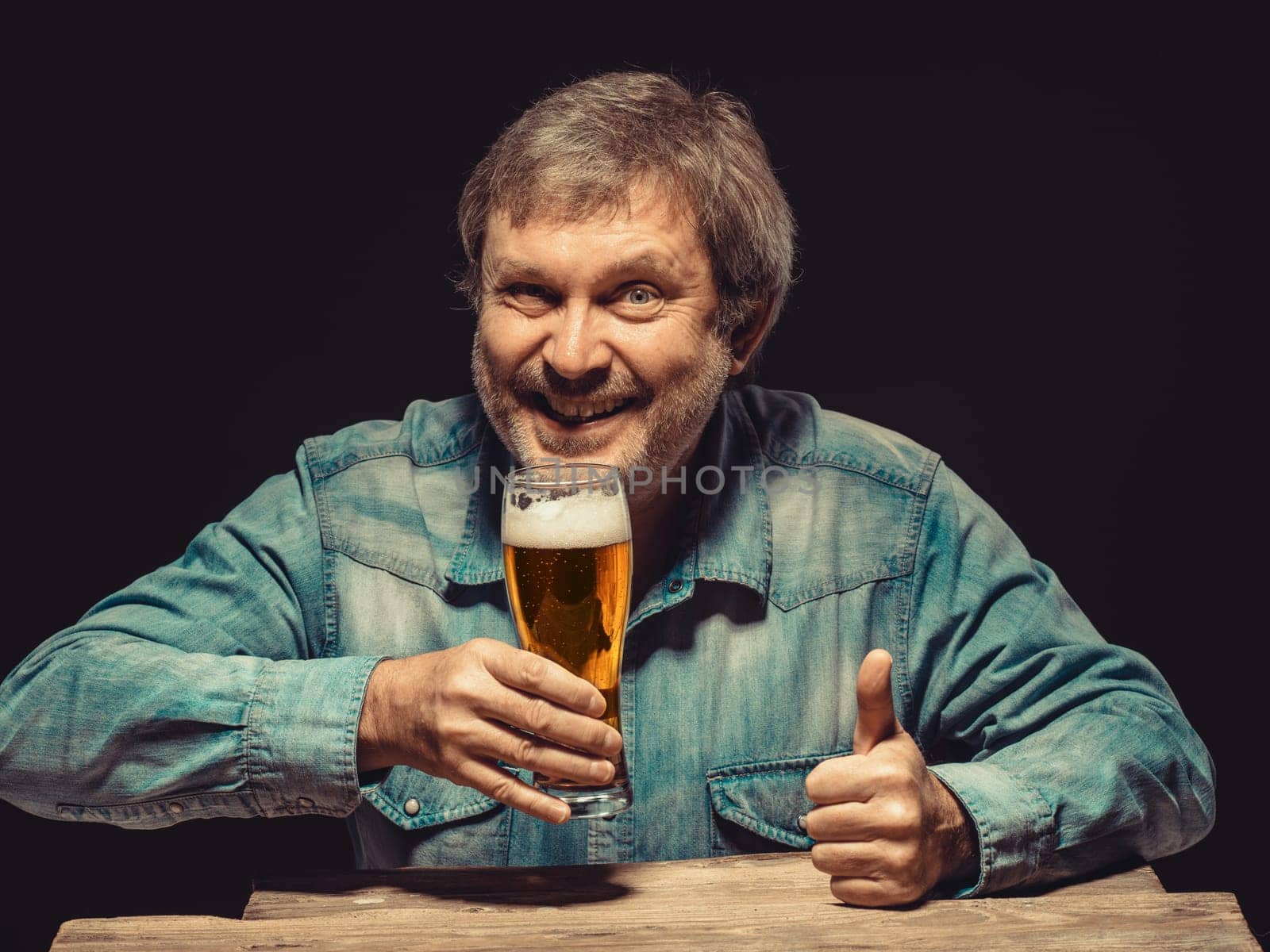Enjoying his favorite beer.  The front view of handsome smiling  man as fan in denim shirt with glass of beer, sitting at the wooden table