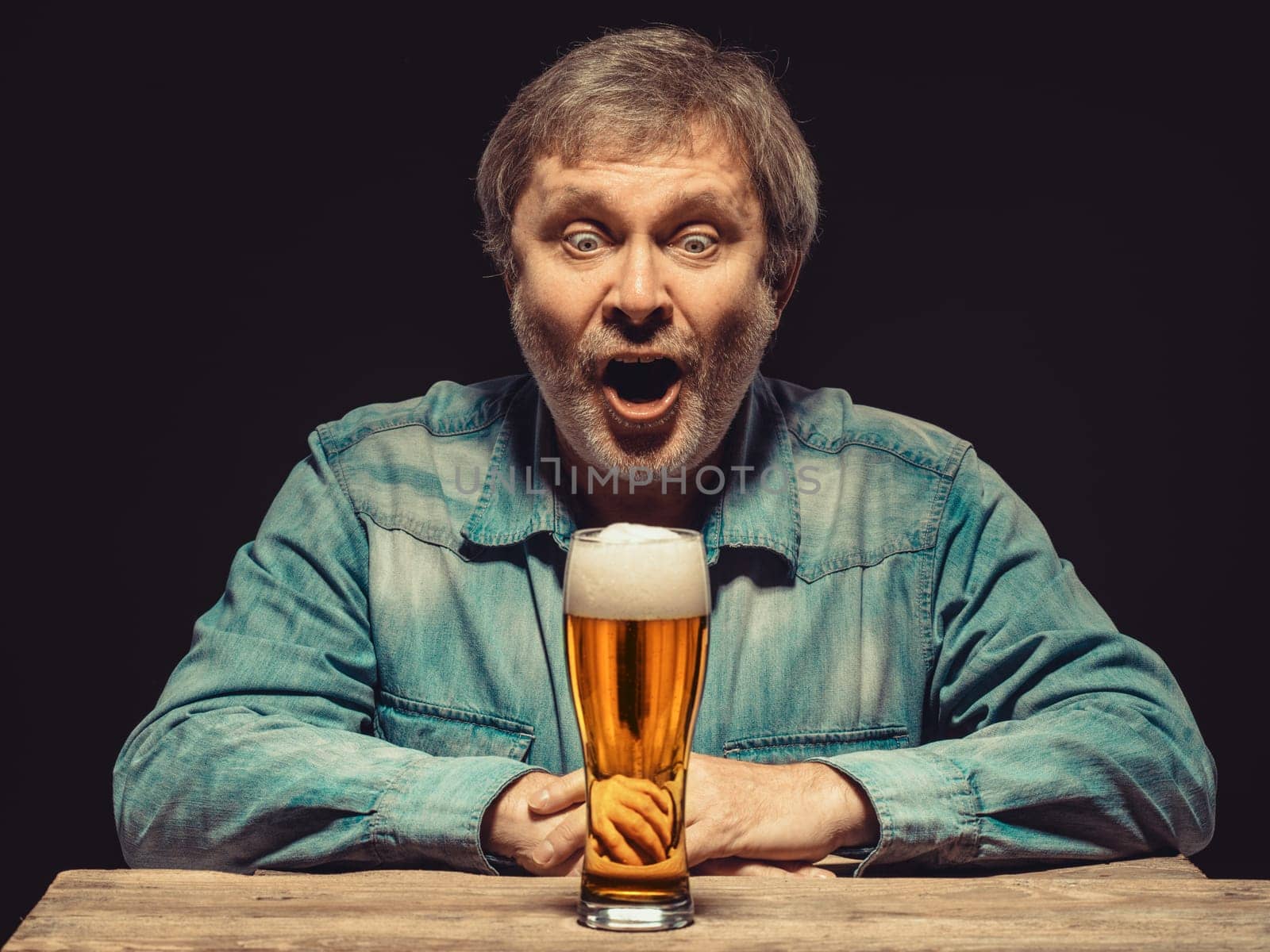 Enjoying his favorite beer.  The front view of handsome screaming man as fan in denim shirt with glass of beer, sitting at the wooden table. Concept of enthusiasm and ecstasy