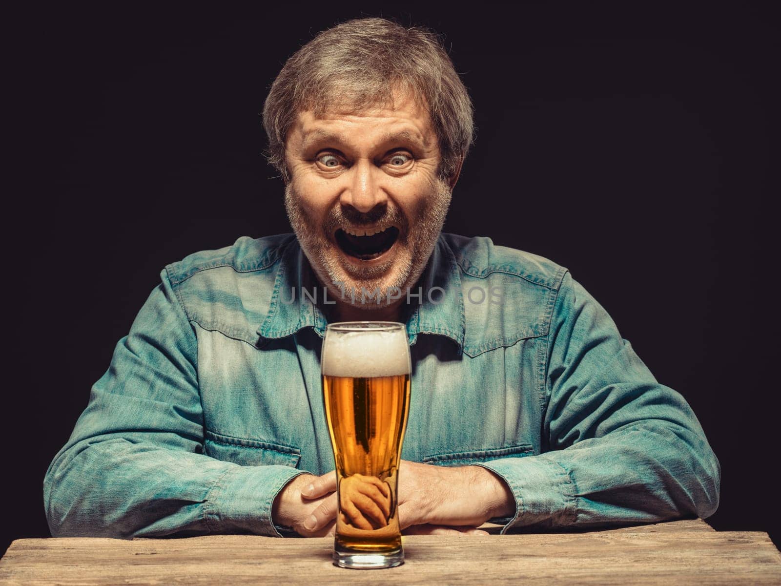 Enjoying his favorite beer.  The front view of handsome smiling  man as fan in denim shirt with glass of beer, sitting at the wooden table. Concept of enthusiasm and ecstasy