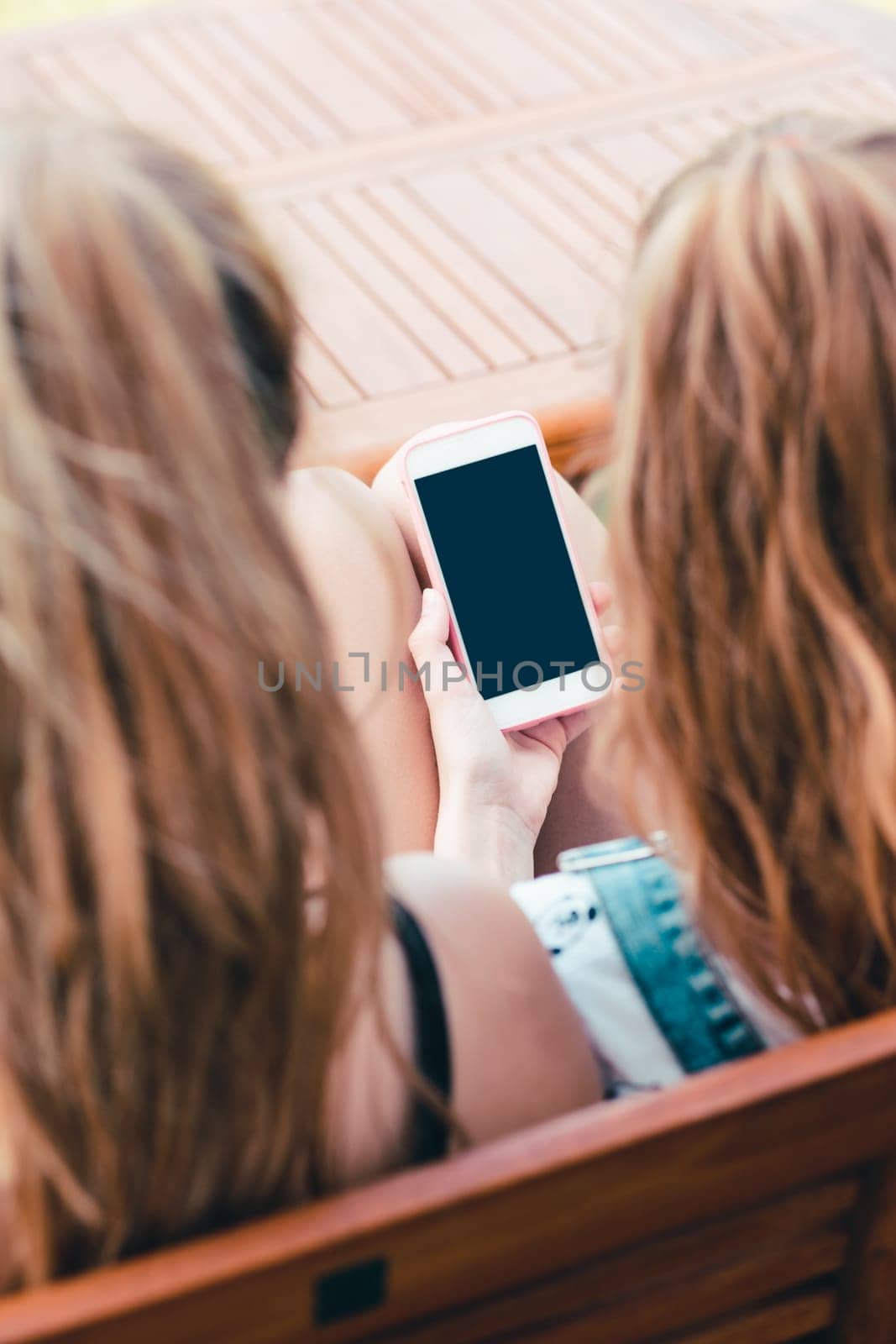 Young women using mobile phones watching music clip, texting, messaging. Teenagers using the smartphones, sitting on bench outdoors. Using technology devices. Girls wearing summer clothing