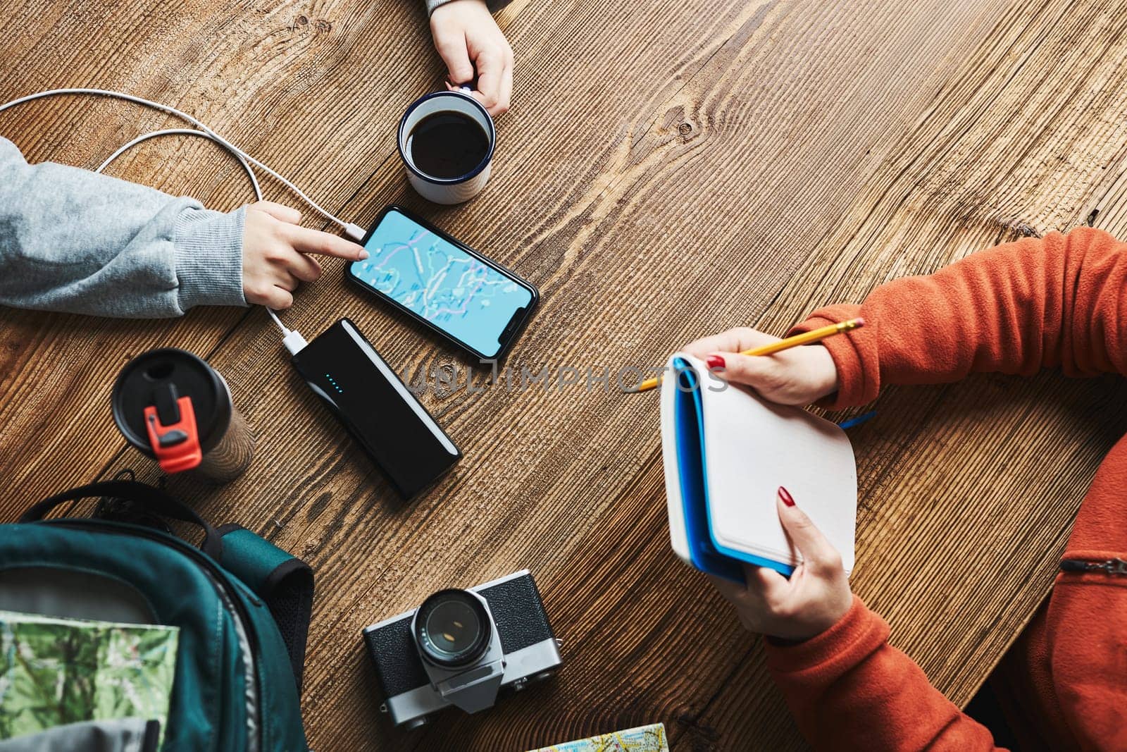Family planning vacation trip. Girl searching travel destination and routes using navigation map on mobile phone. Woman writing journal and making notes. Charging smartphone with power bank. Using technology while travelling. People sitting at wooden table with camera and backpack during journey. Travel plan concept