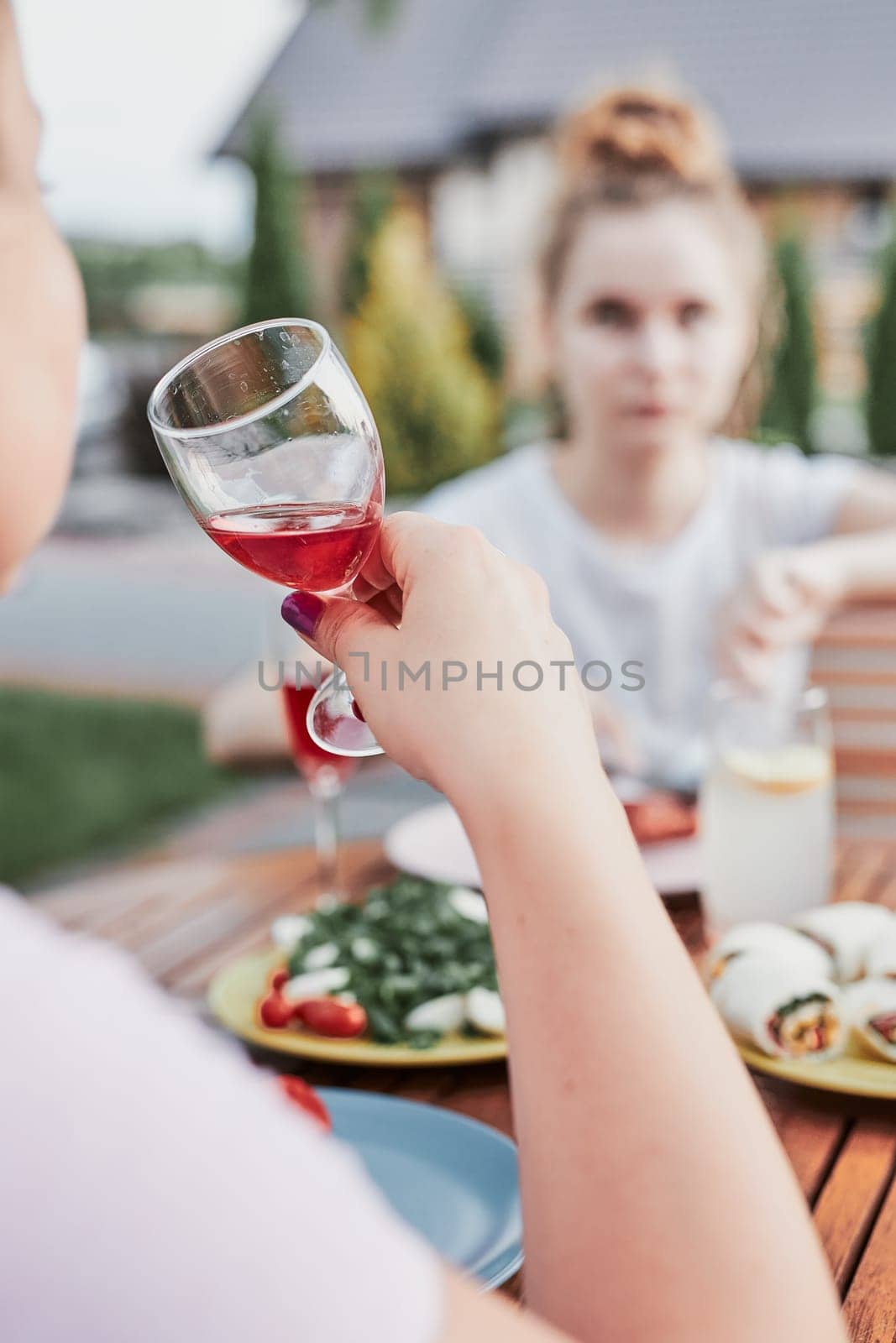 Woman drinking red wine during summer outdoor dinner in a home garden. Close up of hand holding wine glass with red wine over table with dishes