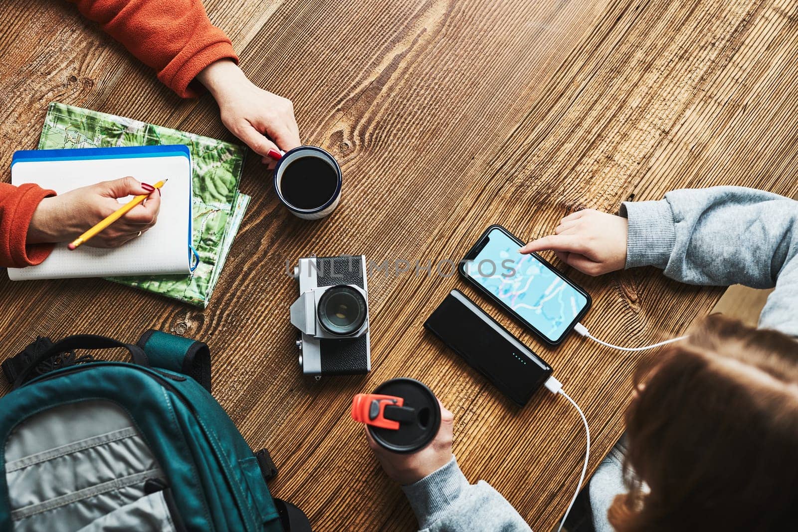 Family planning vacation trip. Girl searching travel destination and routes using navigation map on mobile phone. Woman writing journal and making notes. Charging smartphone with power bank. Using technology while travelling. People sitting at wooden table with camera and backpack during journey. Travel plan concept