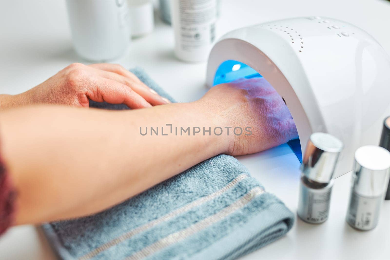 Woman applying gel hybrid polish using UV lamp. Beauty wellness spa treatment concept. Cosmetic products, UV lamp, green leaves on white table. Spa, manicure, skin care concept. Flat lay, overhead view