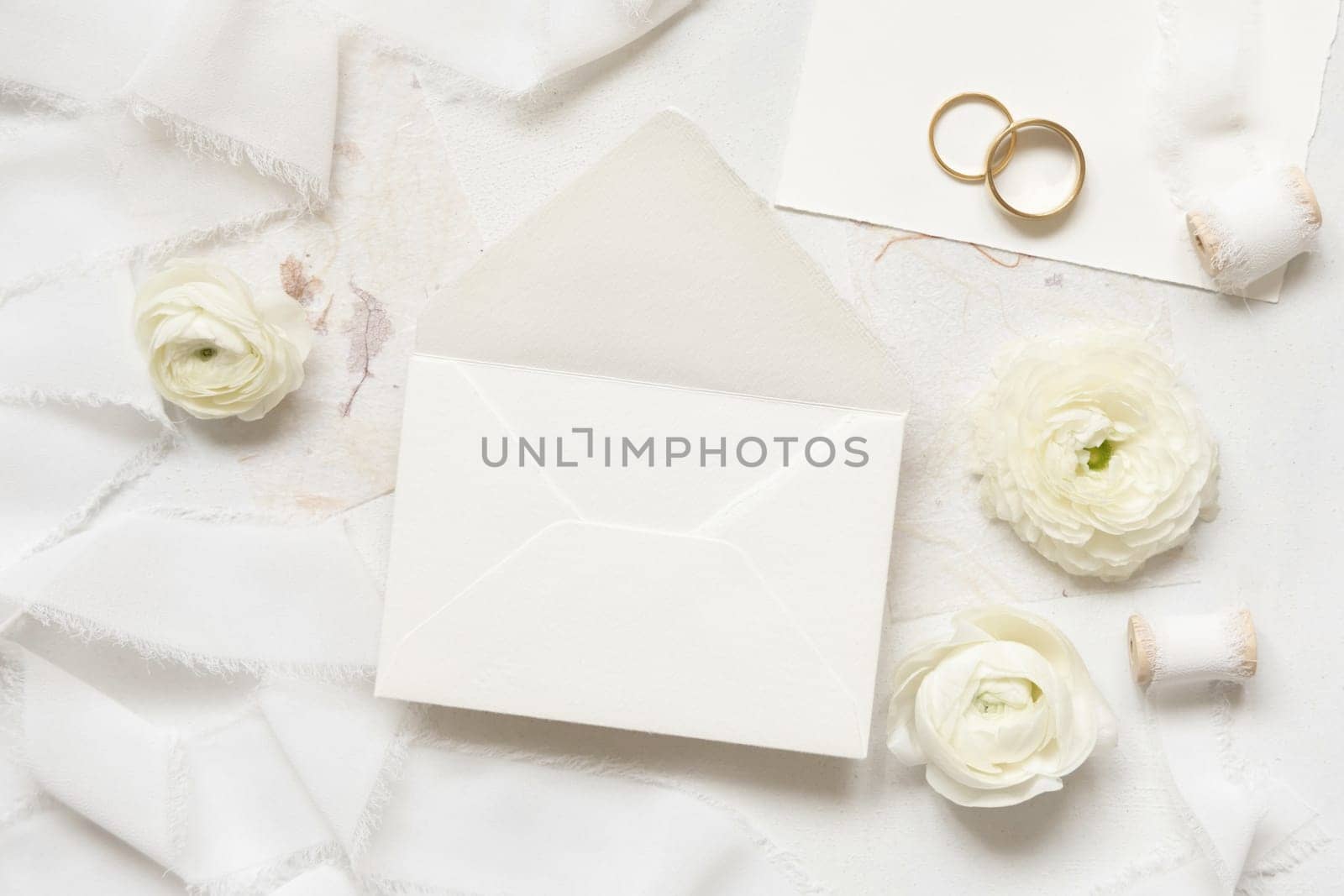 Blank envelope near cream roses, white silk ribbons and wedding rings top view, wedding mockup. Romantic scene with pastel flowers flat lay. Valentines, Spring or Mothers day concept