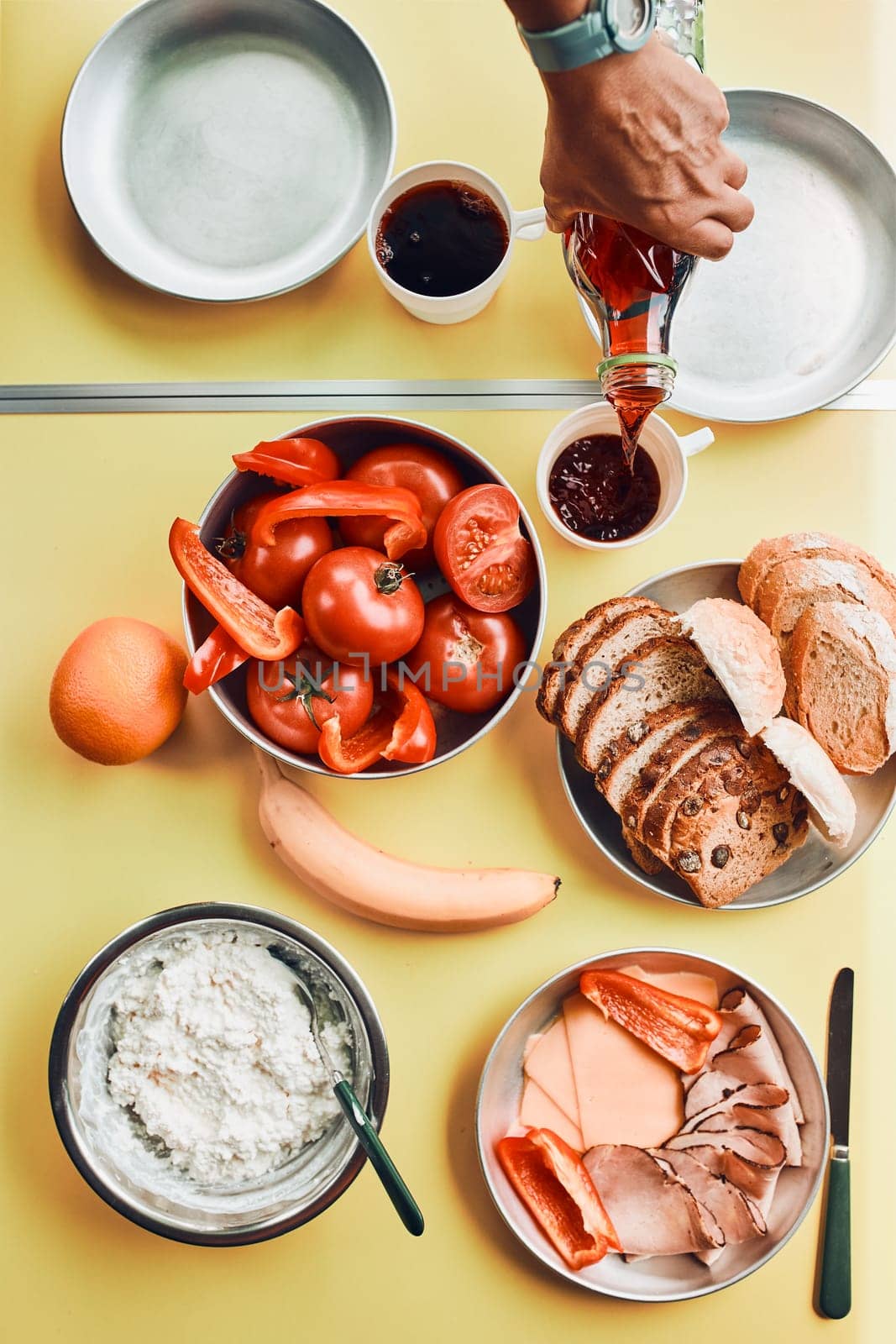 Breakfast prepared during summer vacation on camping. Bread, cottage cheese, cold meat, tomatoes, fruits and coffee cups on table. Close up of outdoor table setting from above