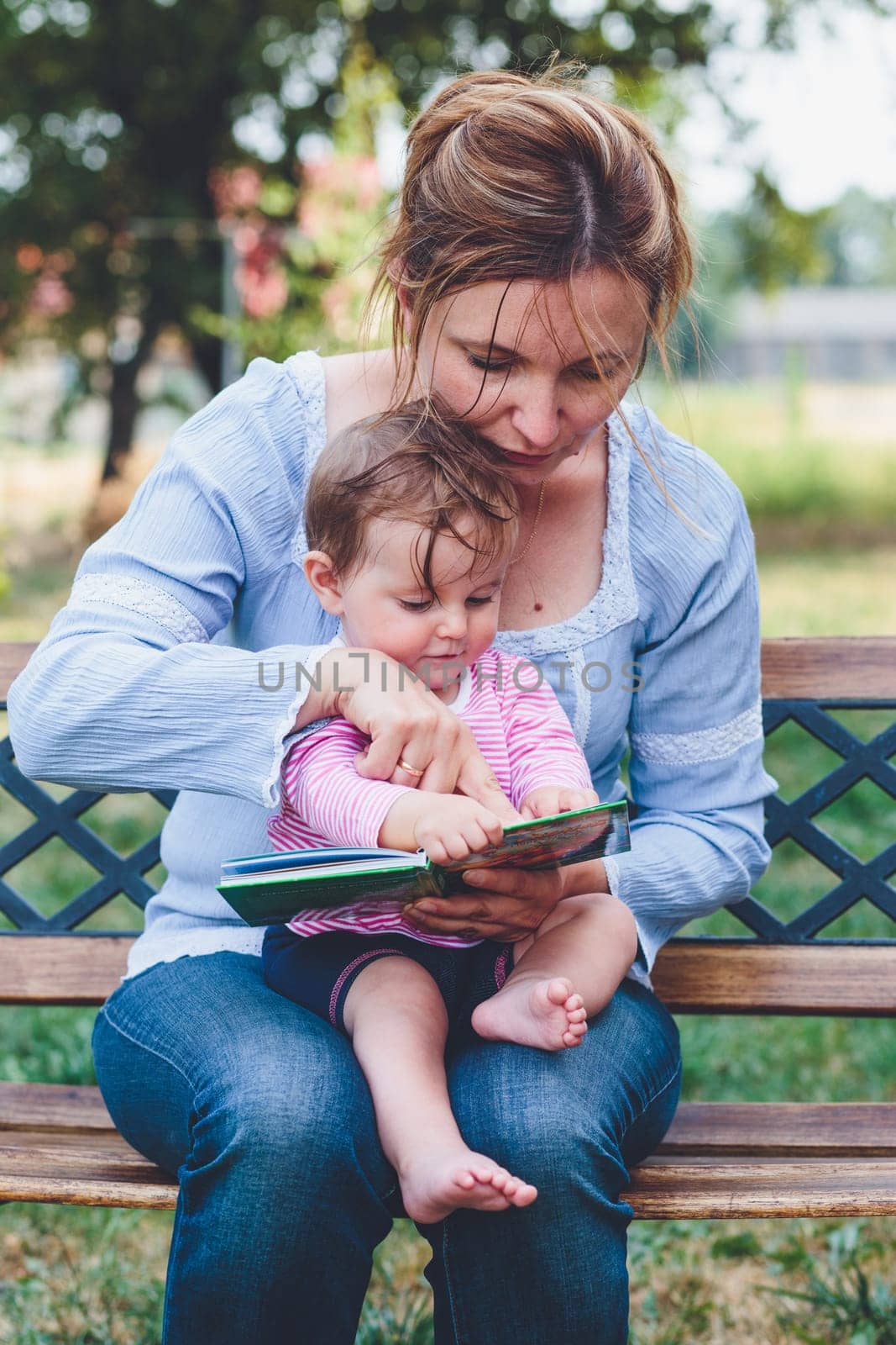 Mom reading a book and showing the pictures her little daughter sitting on bench in the garden