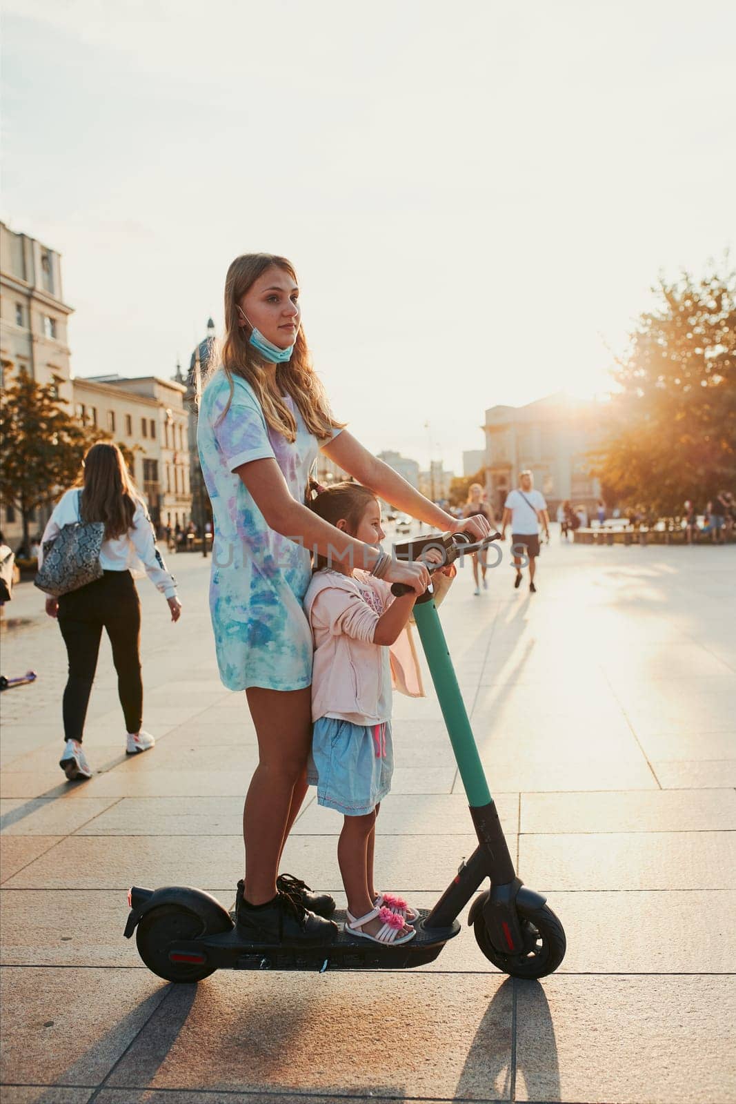 Teenager girl and her sister preschooler riding an electric scooter in the city center. Concept of happy family. Candid people, real moments, authentic situation