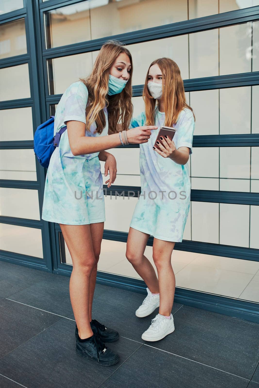 Young women girls using smartphones using social media sending texts watching photos and videos. Friends spending time talking with friends standing at store front downtown. Girls wearing the face masks to avoid virus infection and to prevent the spread of disease in time of coronavirus