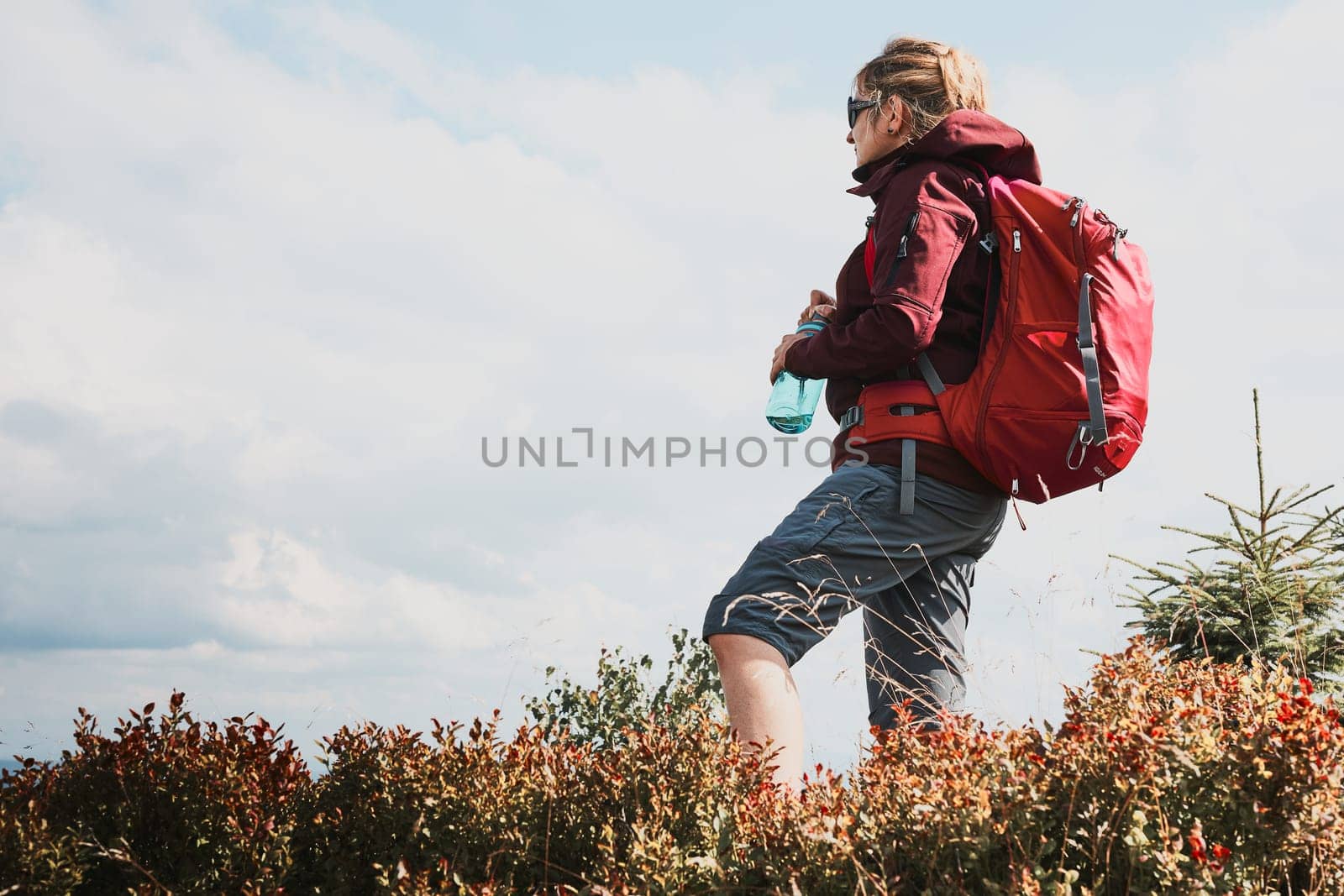Woman with backpack hiking in mountains, spending summer vacation close to nature. Woman standing on top of hill admiring mountain landscape panorama holding bottle with water